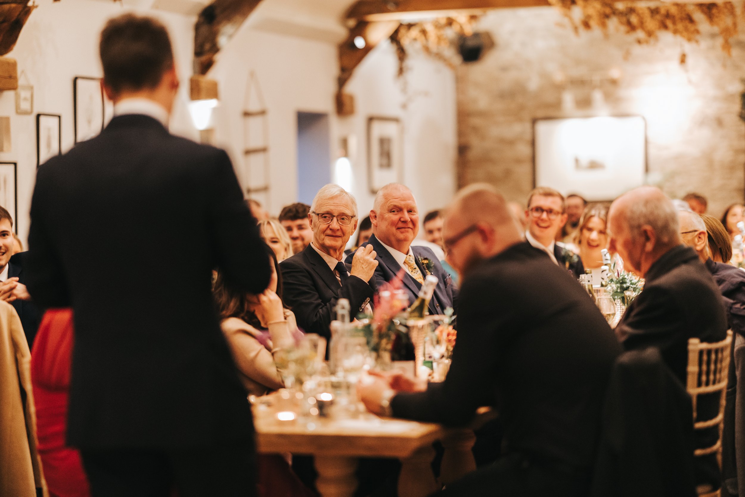 People sitting at a banquet table during a formal event, with a man standing and speaking to the group in a warmly lit room with wood beams and framed pictures on the walls.