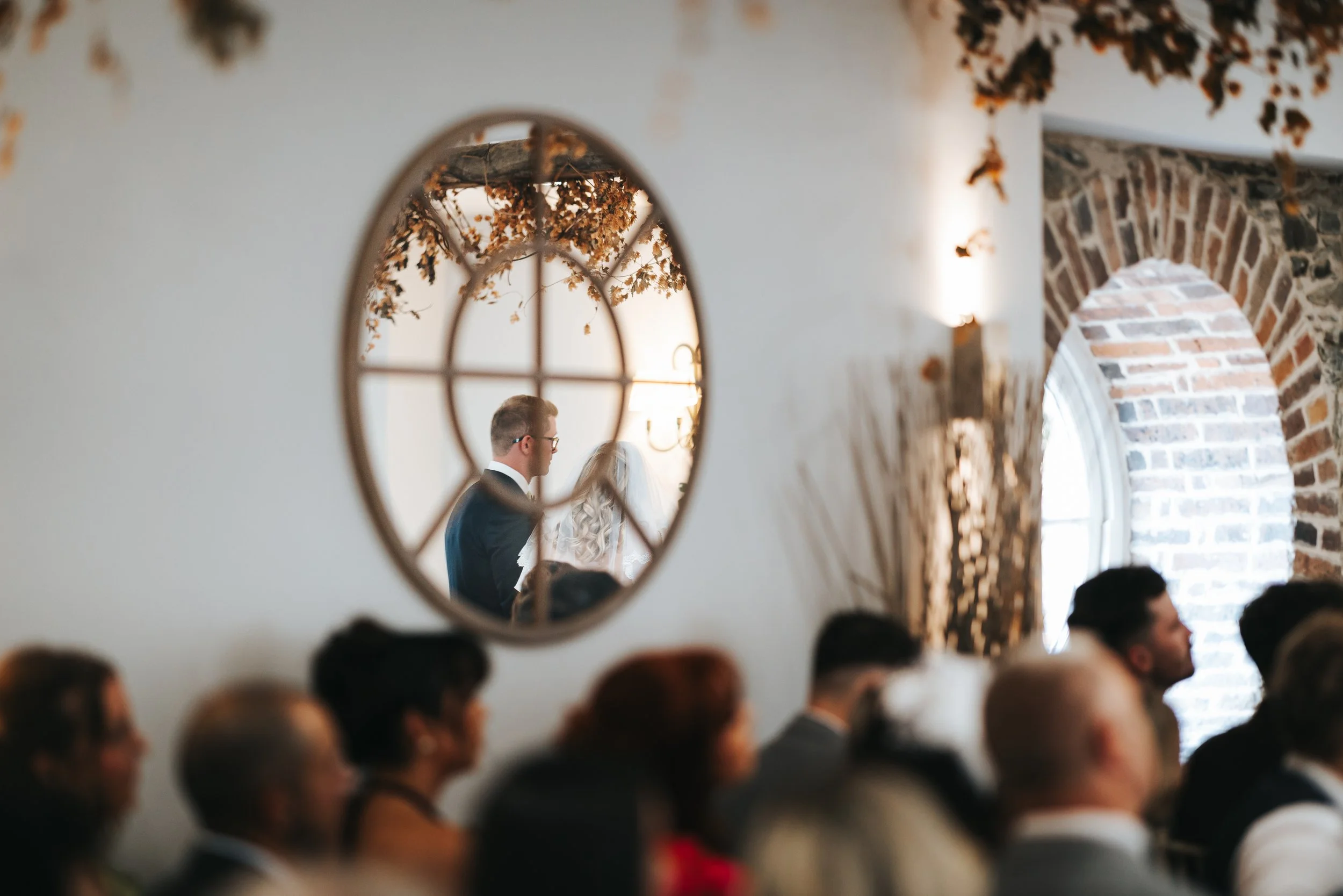 A wedding ceremony seen through an oval mirror on a white wall, reflecting a bride and groom. The groom has glasses and a black tuxedo, and the bride has long blonde hair. Guests are seated in front, with brick walls and large arched windows in the b