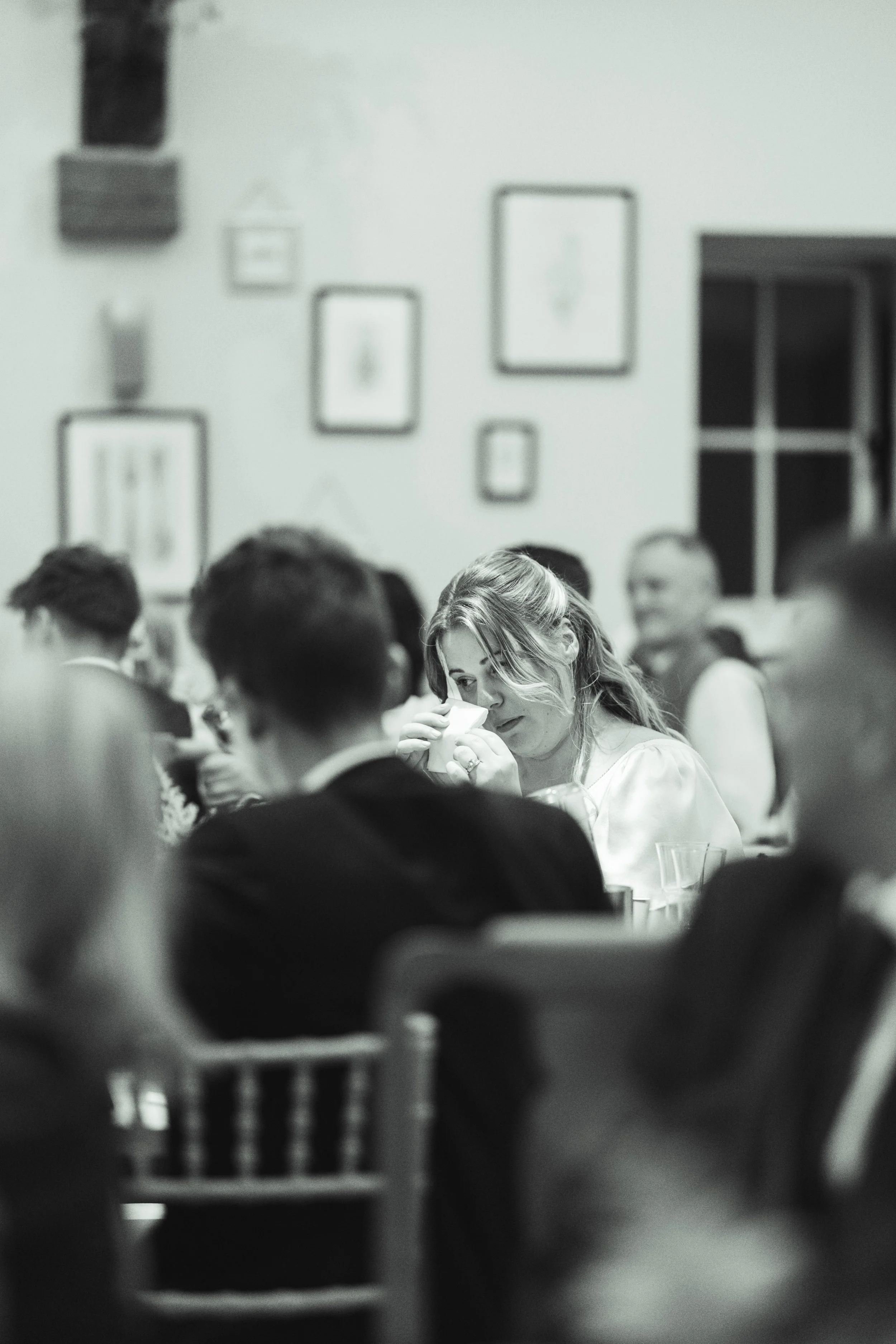 A black and white photo of a woman at a gathering, likely crying or emotional, holding a tissue to her face, surrounded by other people in a room with framed pictures on the wall.