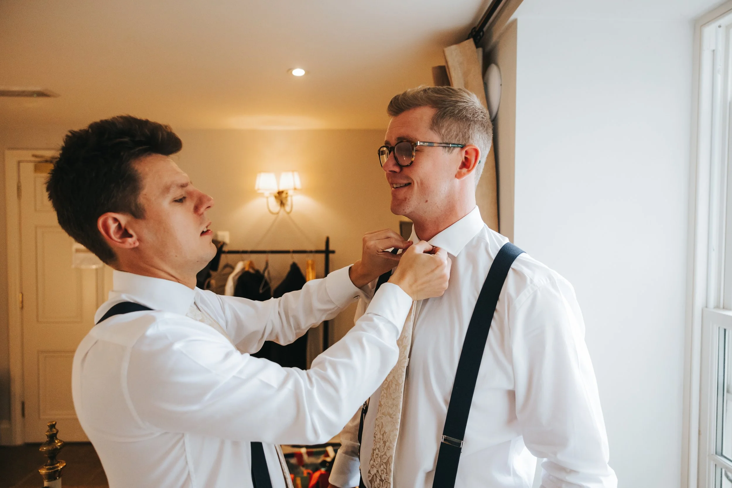 Two men in white dress shirts, black suspenders, and a tie, are getting ready together in a room. One is adjusting the other's tie, and they are smiling. The room has warm lighting, a window, and some clothing and hangers in the background.