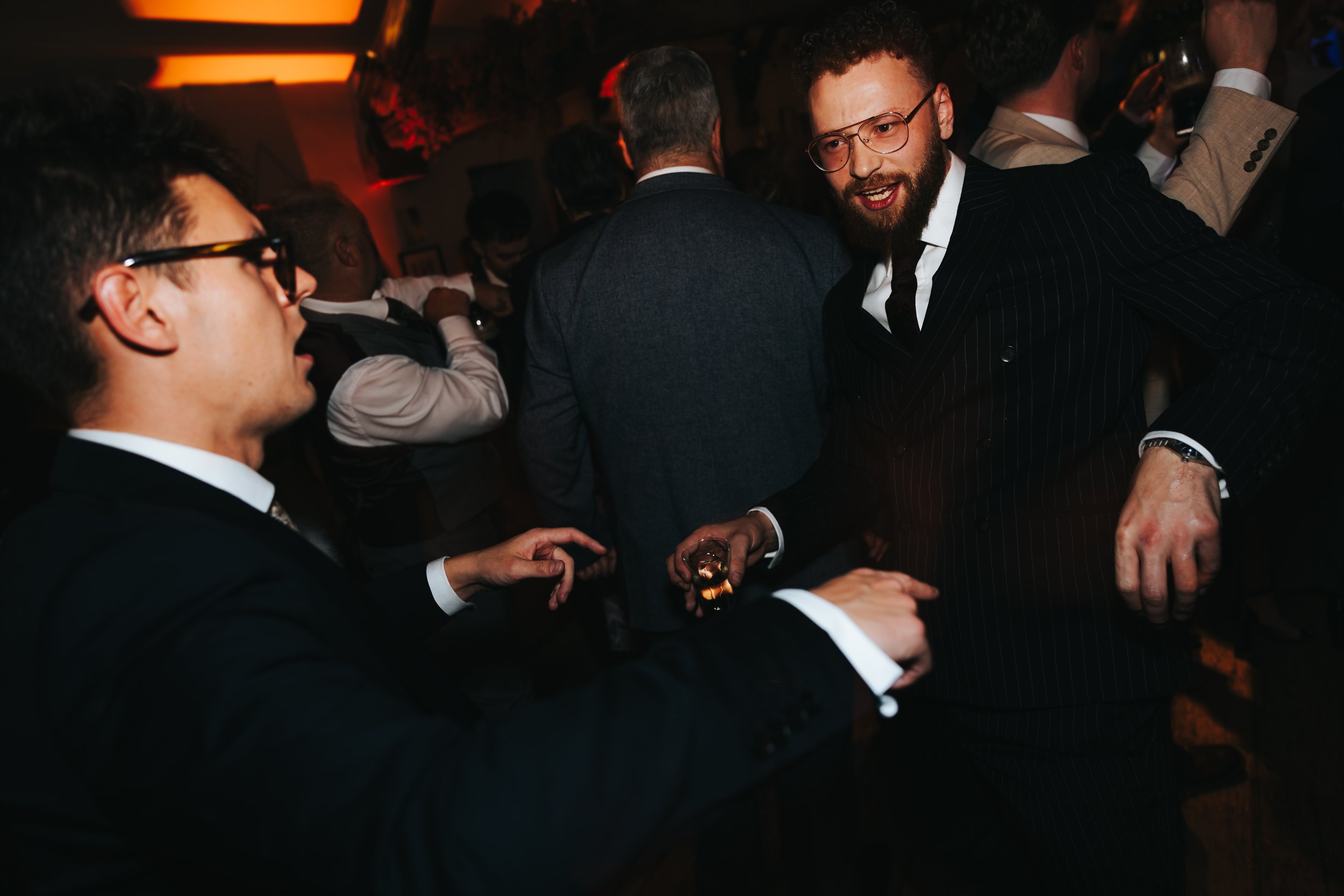 Two men in suits dancing and chatting at a party, with other people in the background.