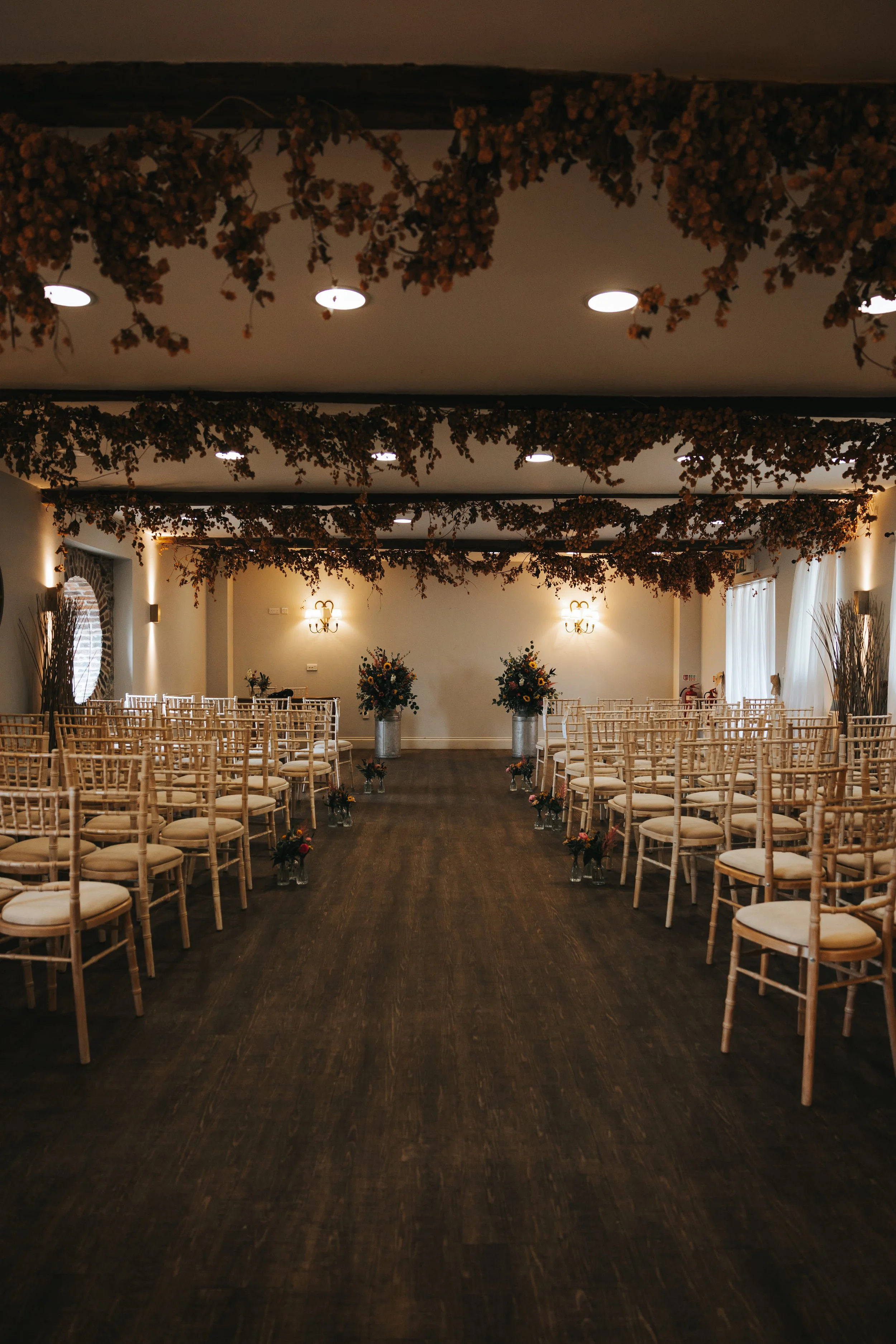 A decorated wedding ceremony room with rows of wooden chairs and floral arrangements, hanging dried flowers on the ceiling, and soft lighting.