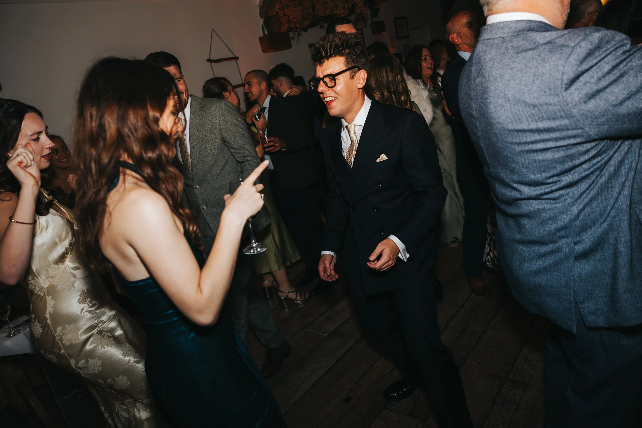 People dancing and enjoying at a lively party or wedding celebration, dressed in formal attire, with a man in a suit and glasses dancing in the center.