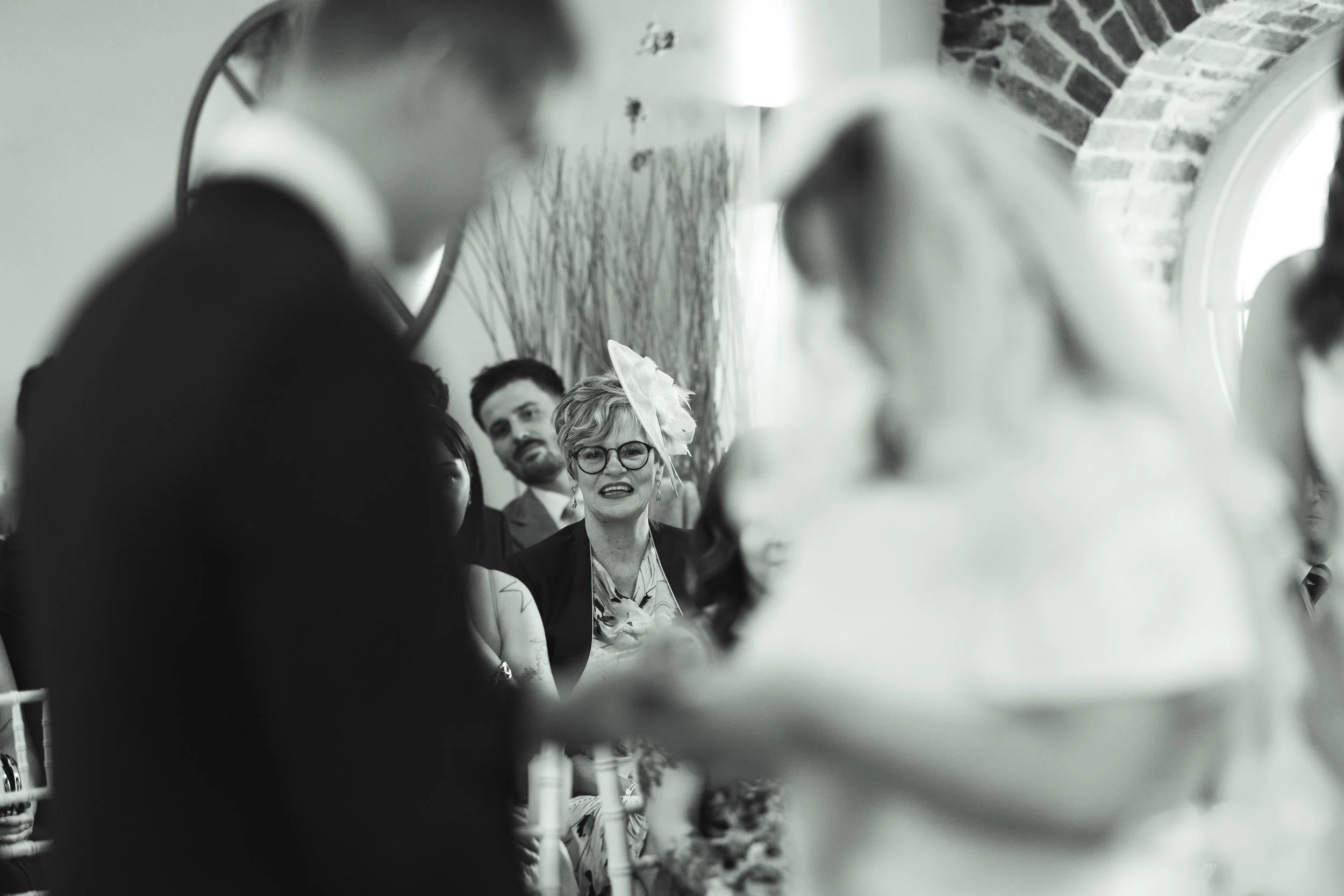 Black and white photo of a woman with glasses and a hat smiling at a gathering, seen through a mirror with several people around her.