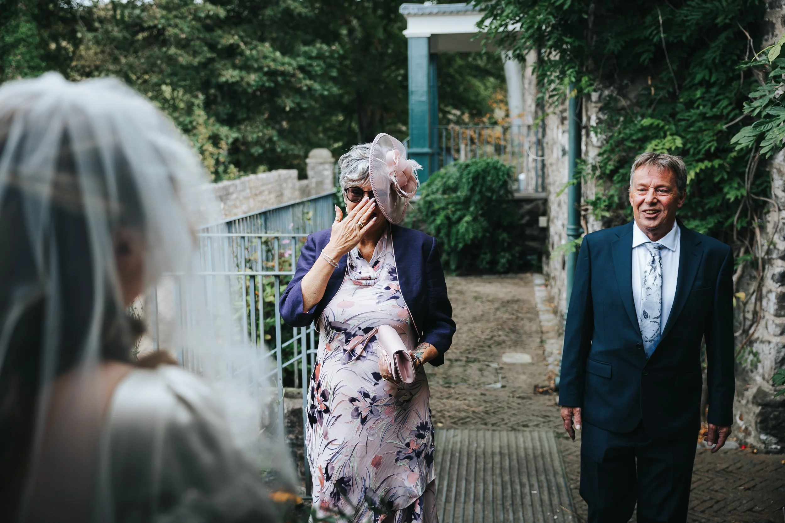 A woman with short gray hair wearing a large pink hat and sunglasses covers her mouth with her hand, standing outside on a stone pathway next to a man in a dark suit, white shirt, and patterned tie. Another woman with a veil is partially visible in t