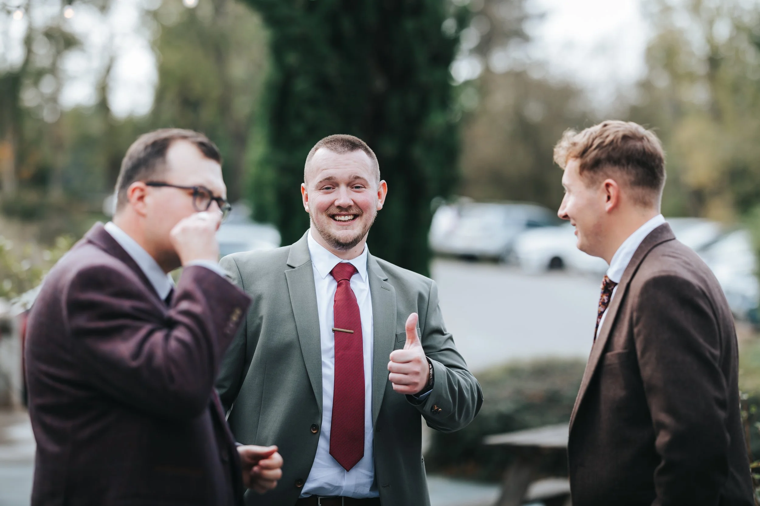 Three men in suits are standing outdoors, smiling and engaging in conversation. One man is giving a thumbs-up gesture and another is wiping his eyes, possibly laughing. The background shows trees and parked cars.
