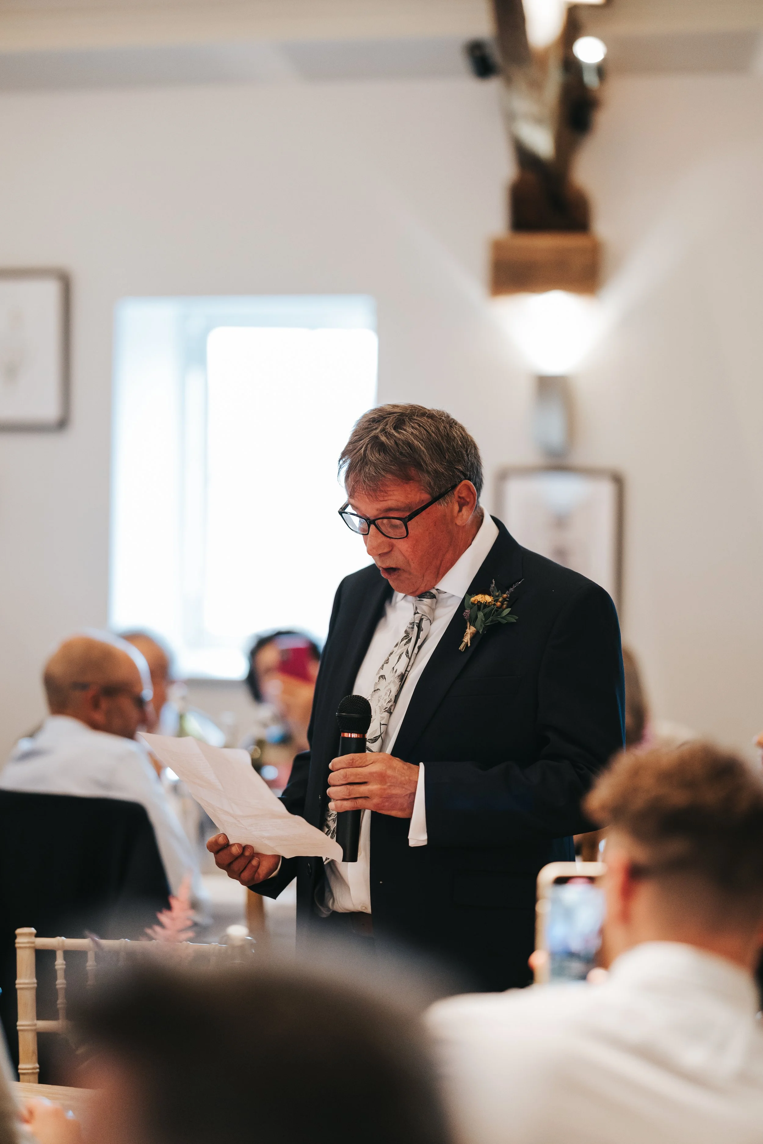 A man in a suit and glasses giving a speech while holding a microphone and reading from a piece of paper at a formal event, with an audience seated in front of him.