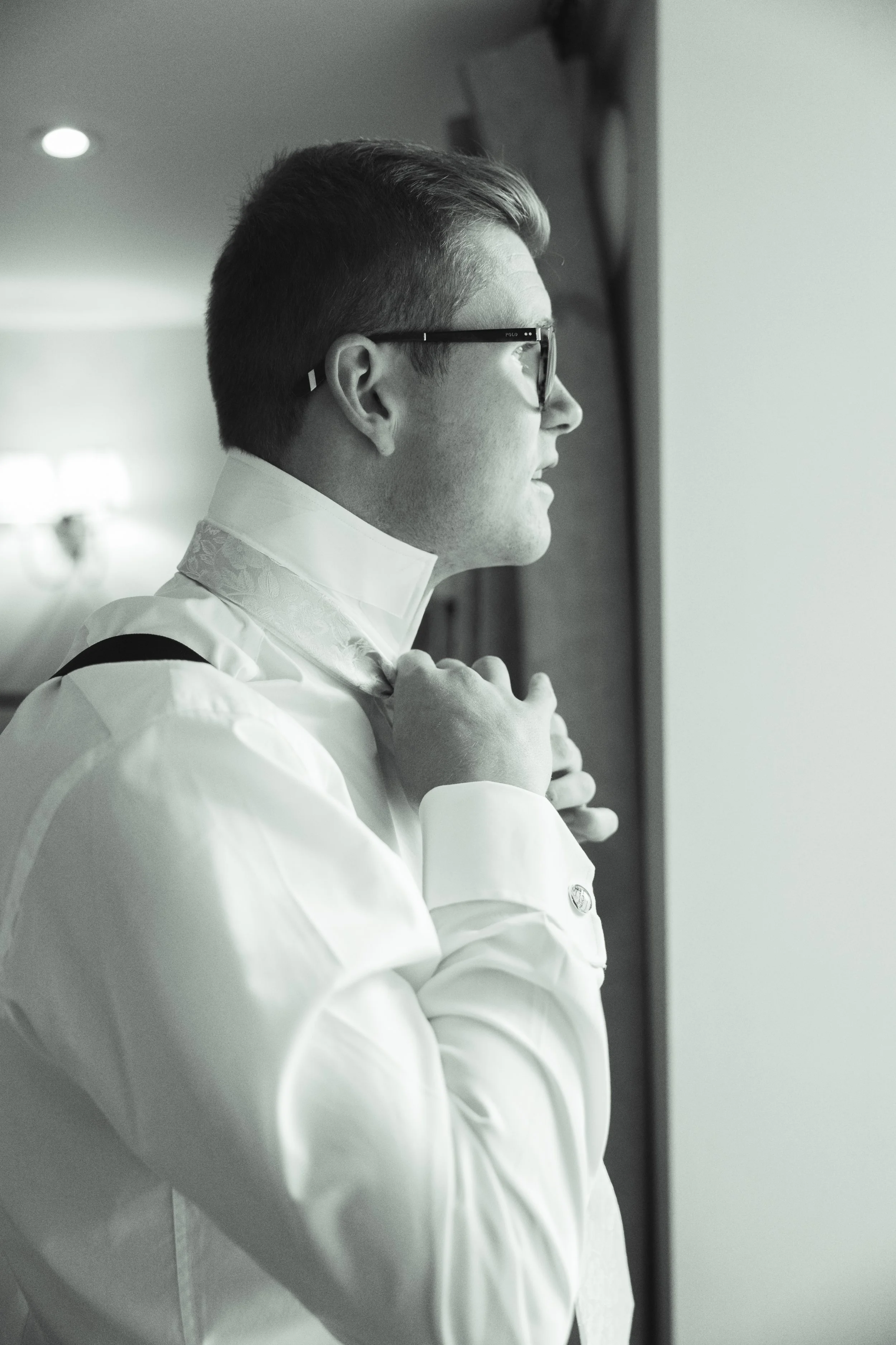A man with glasses adjusts his bow tie while looking out a window.