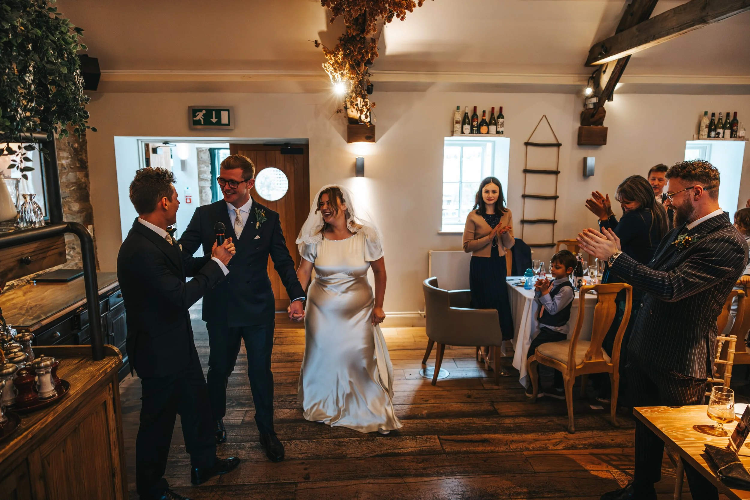 A wedding scene with a bride, groom, and officiant at the center, holding hands. Guests are clapping and smiling in a warmly decorated room with wooden beams and rustic decor.