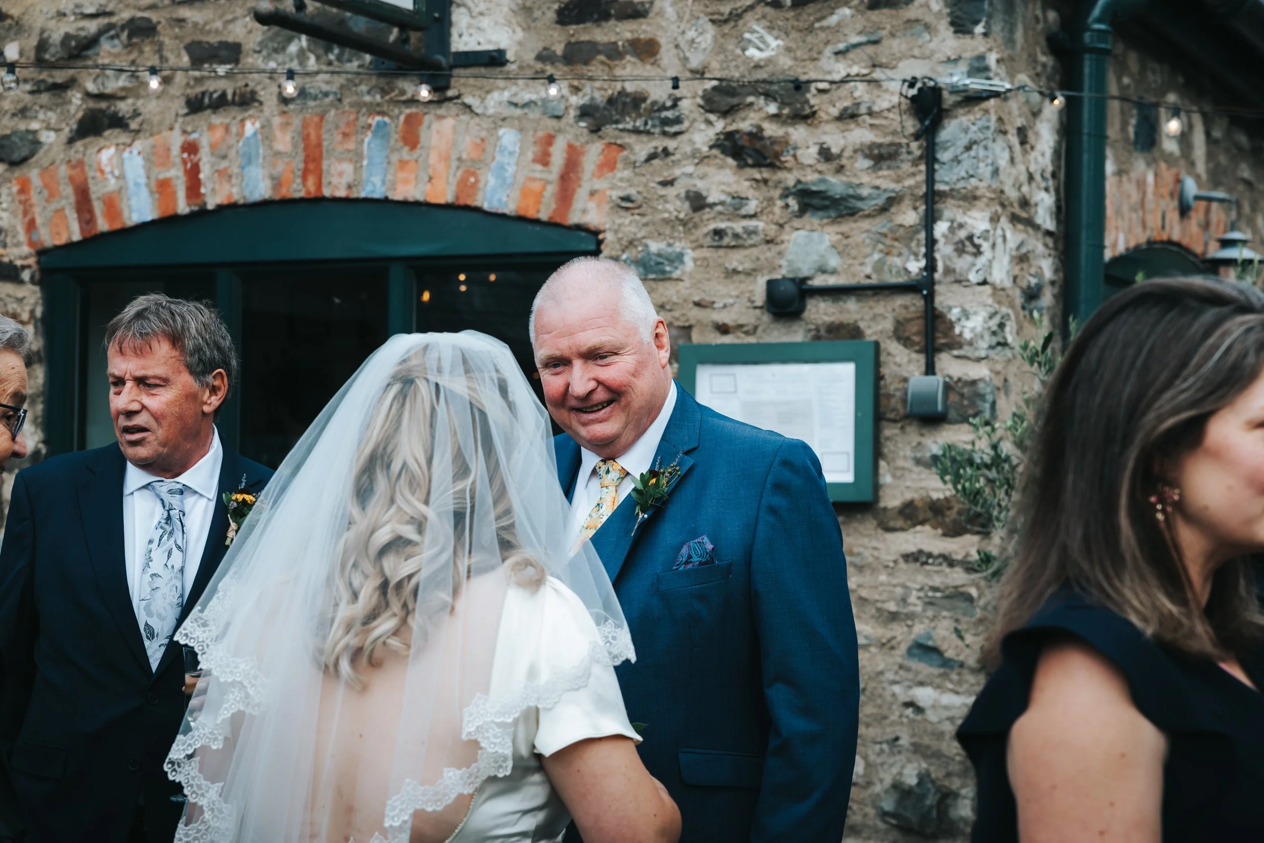 A groom smiling while talking to a bride with a veil during a wedding reception outdoors, with a stone wall in the background.