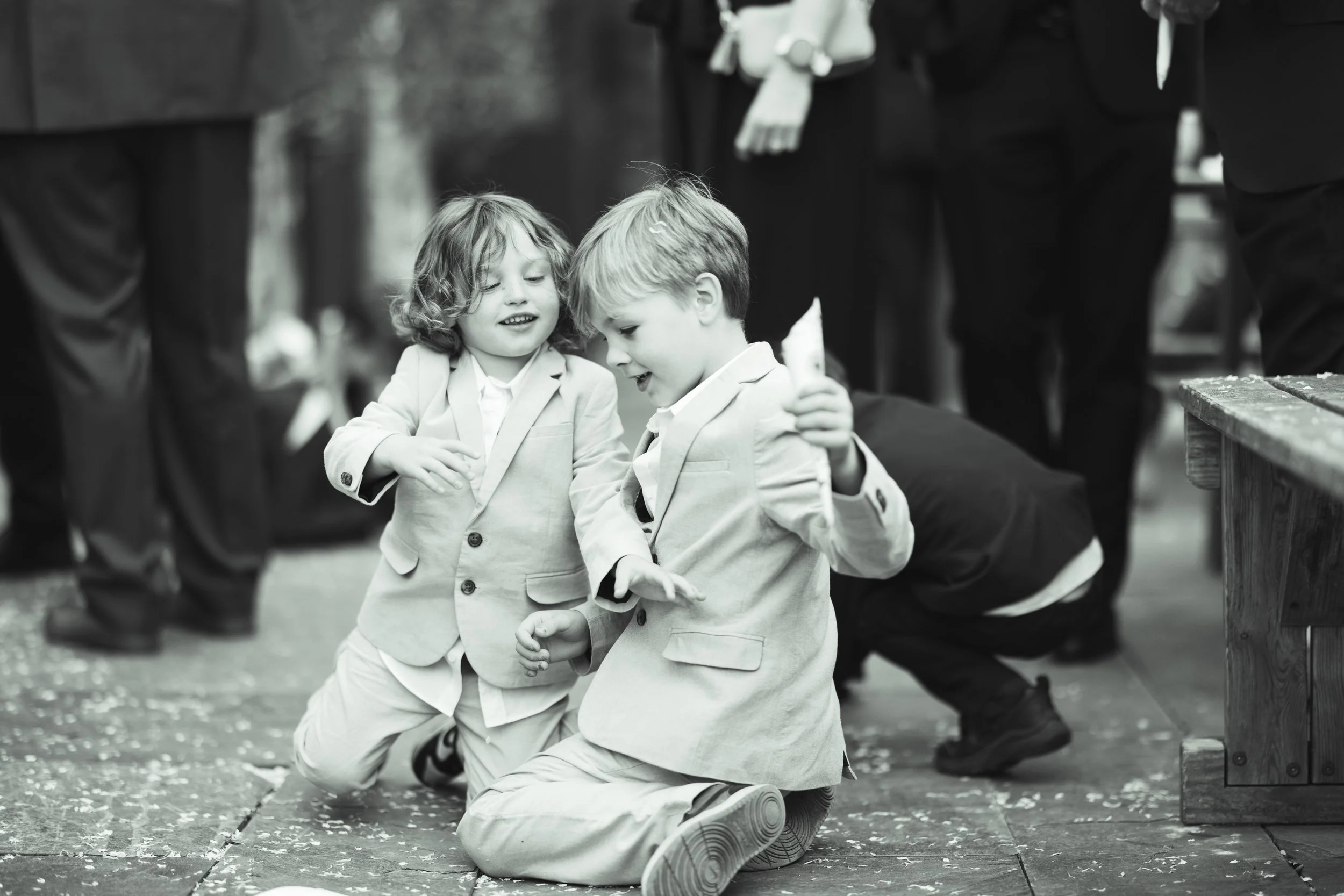 Two young boys in suits kneeling on the ground and smiling, holding a small object, possibly a candy or toy, while other people are visible in the background.