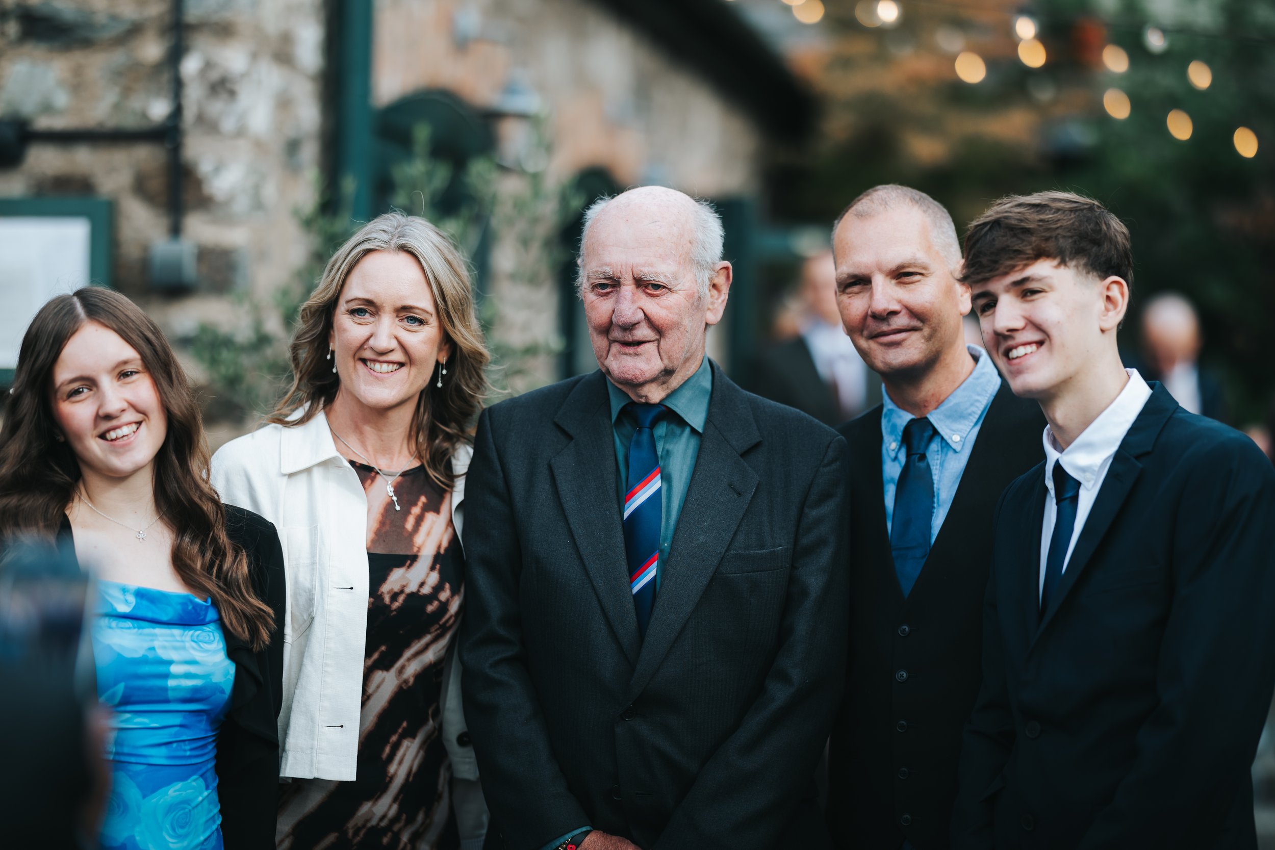 Group of five people, three men and two women, smiling and posing outdoors at a celebration or gathering with blurred background and string lights.