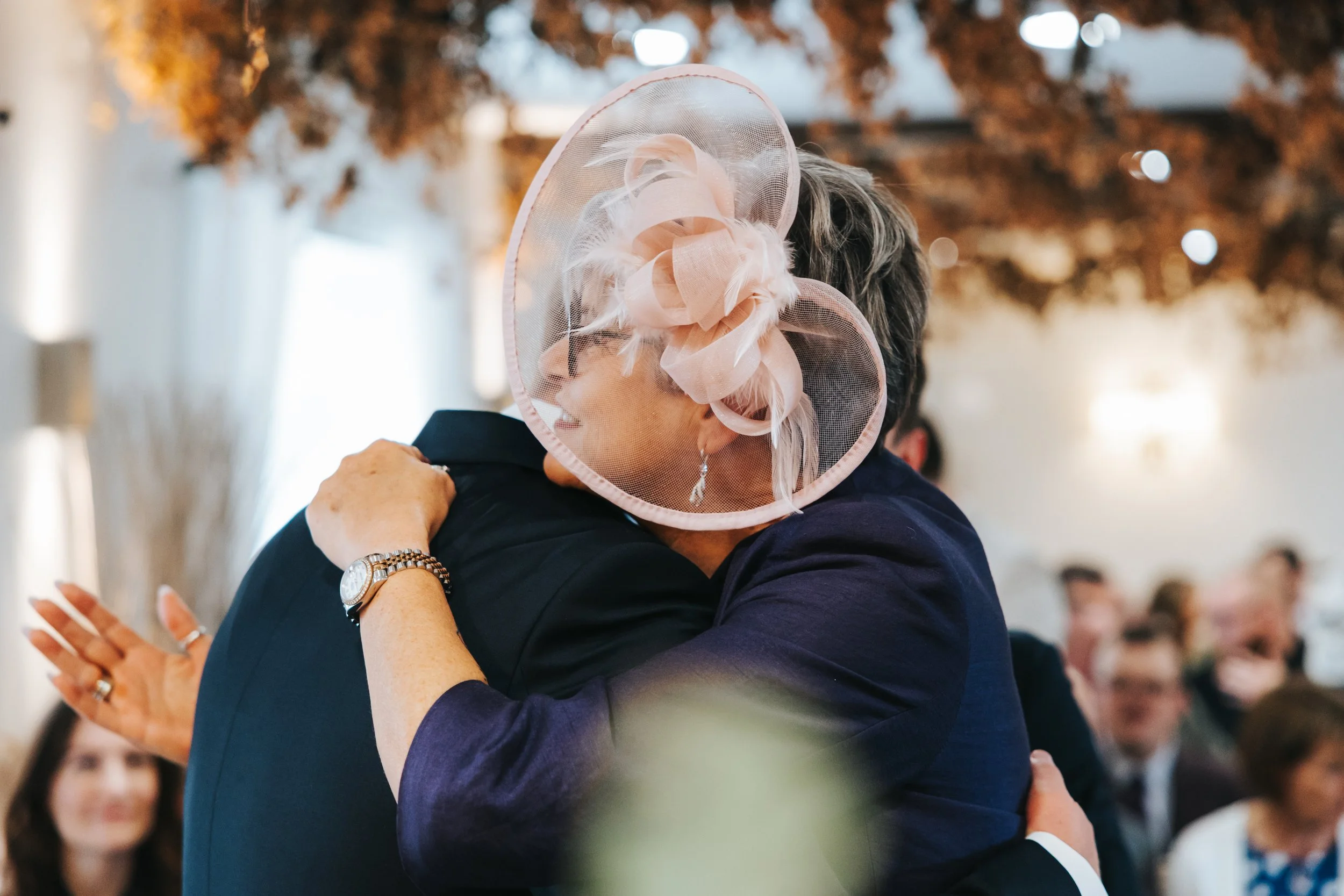 Two women hugging at a ceremony or celebration. One woman wears a large pink fascinator hat with feathers, and the other woman wears a dark blazer. The background shows other people seated, and the setting appears to be decorated for a formal event.