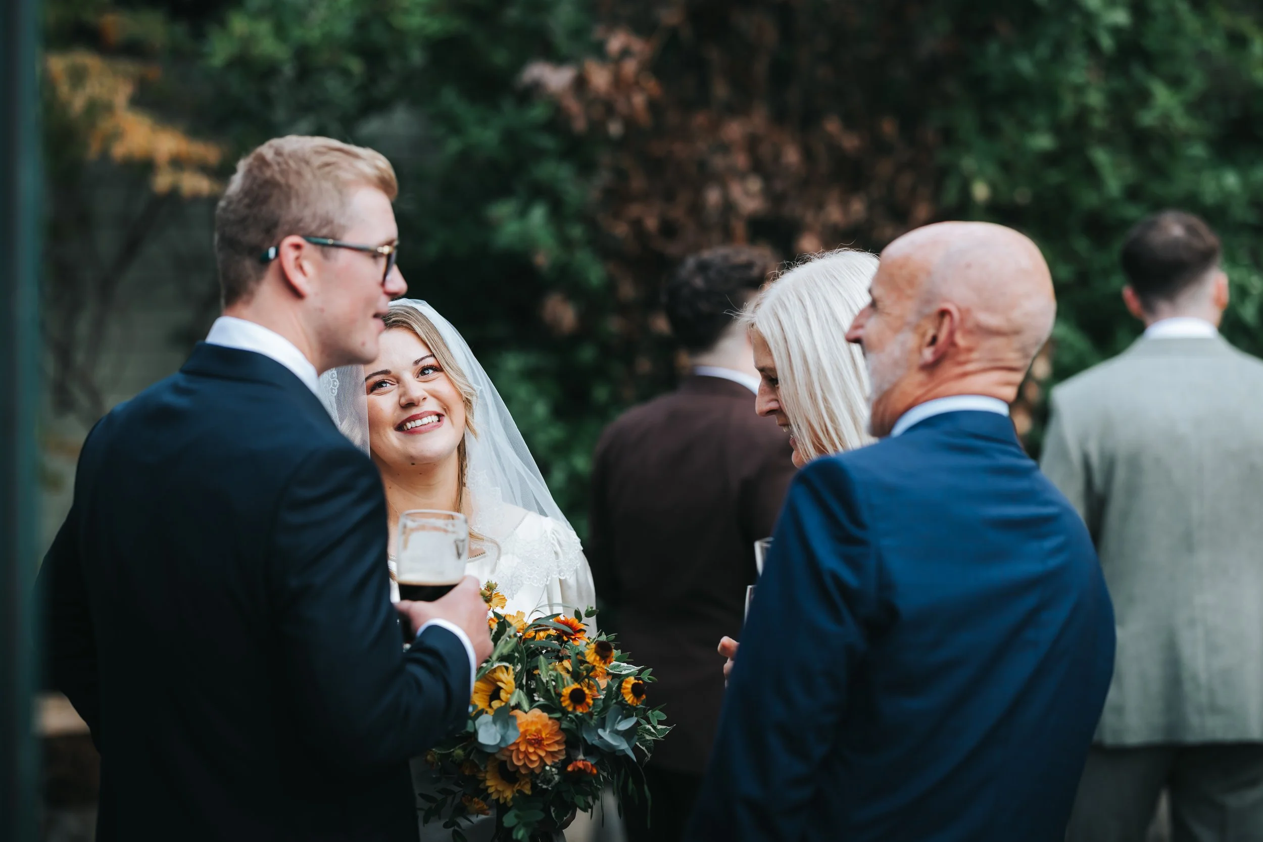 People dressed in formal wear at an outdoor wedding reception socializing, with a woman holding a bouquet of flowers and smiling at a man holding a drink