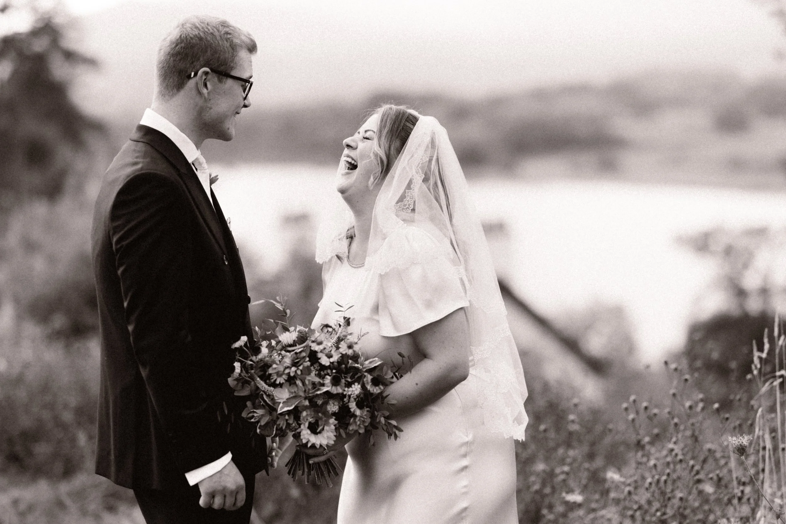 A black-and-white photo of a bride and groom on their wedding day, standing outdoors in a natural setting, with the bride holding a bouquet and laughing while looking at the groom.