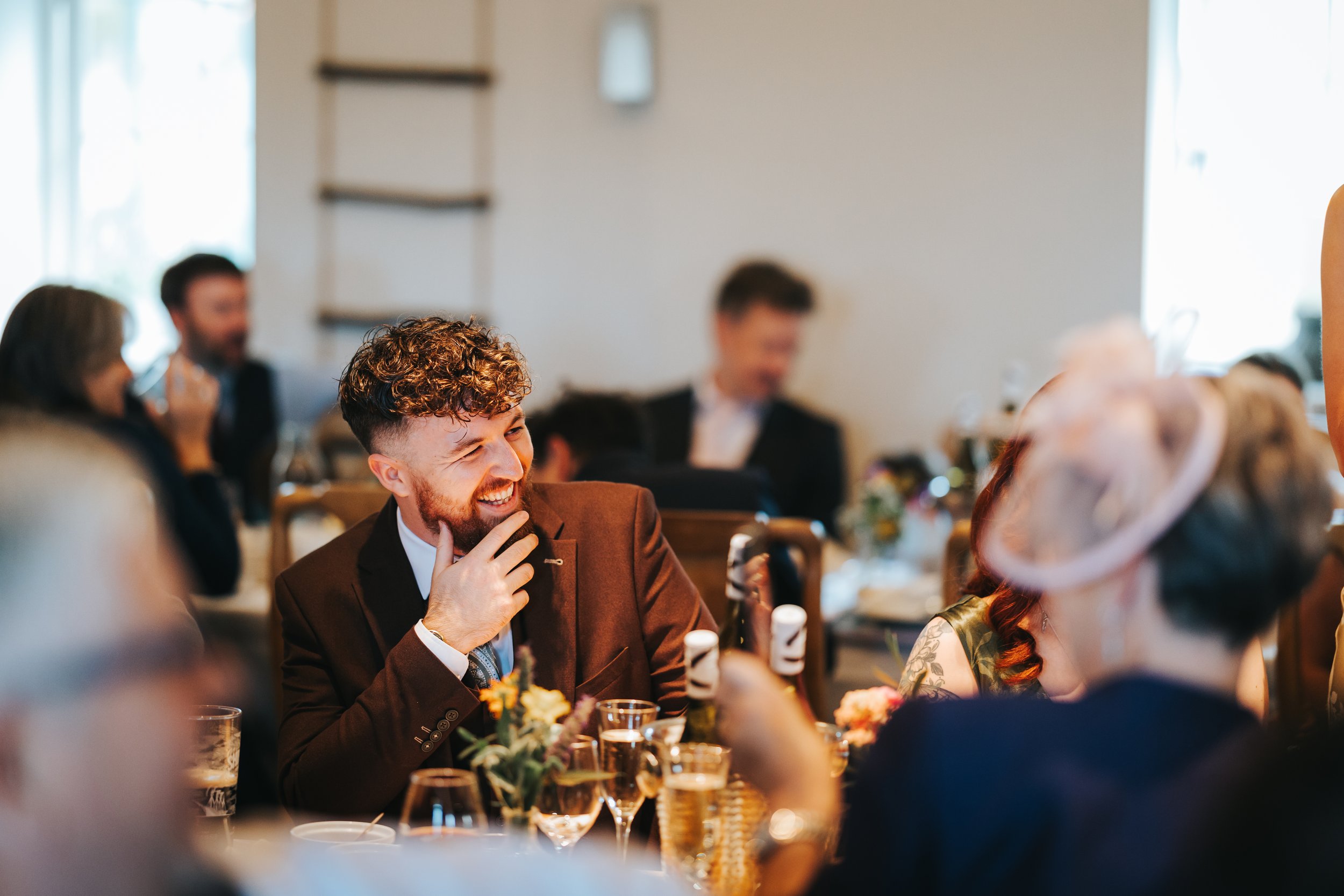 A group of people at a social gathering or celebration, sitting at a table decorated with flowers, drinks, and glasses, with focus on a man with curly hair and a beard wearing a dark suit, smiling and interacting with others.