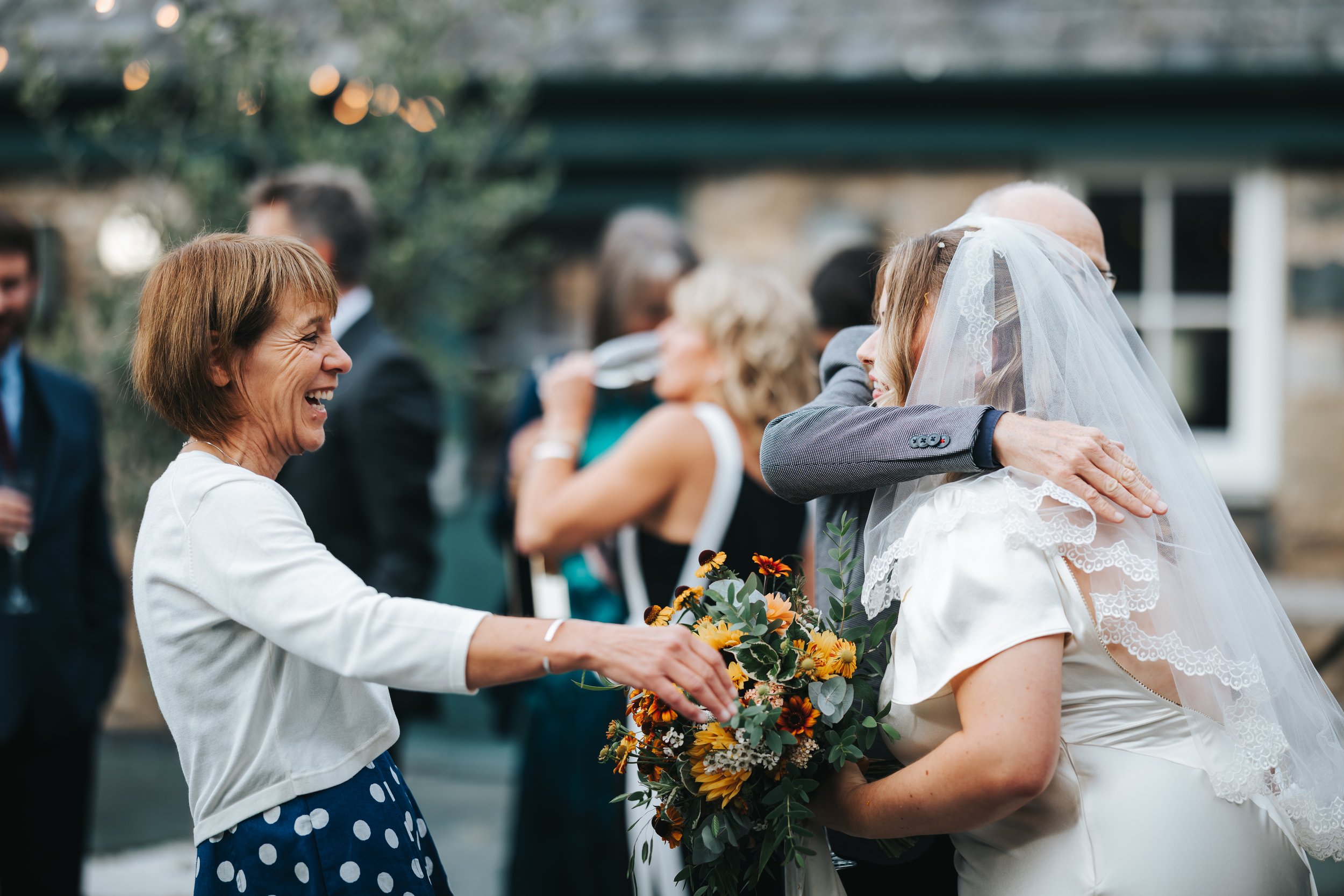 A woman in a white jacket and polka dot dress happily greets a bride in a wedding dress and veil, embracing her while holding a bouquet of flowers at a wedding reception.