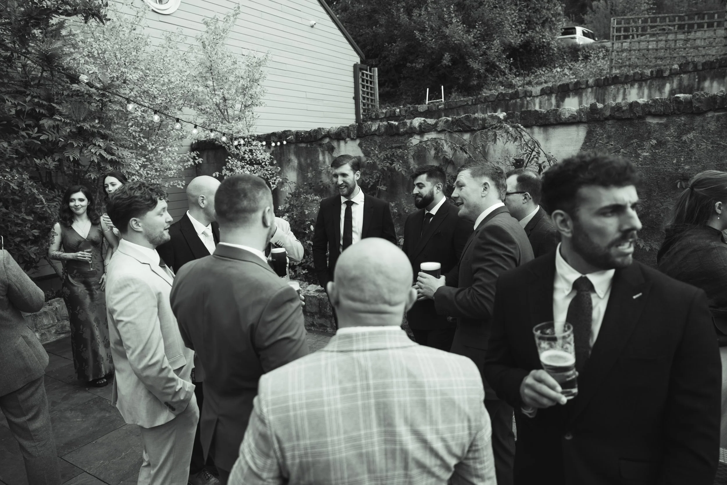 Group of people in formal attire socializing outdoors at night, some holding drinks, with string lights and a garden in the background.