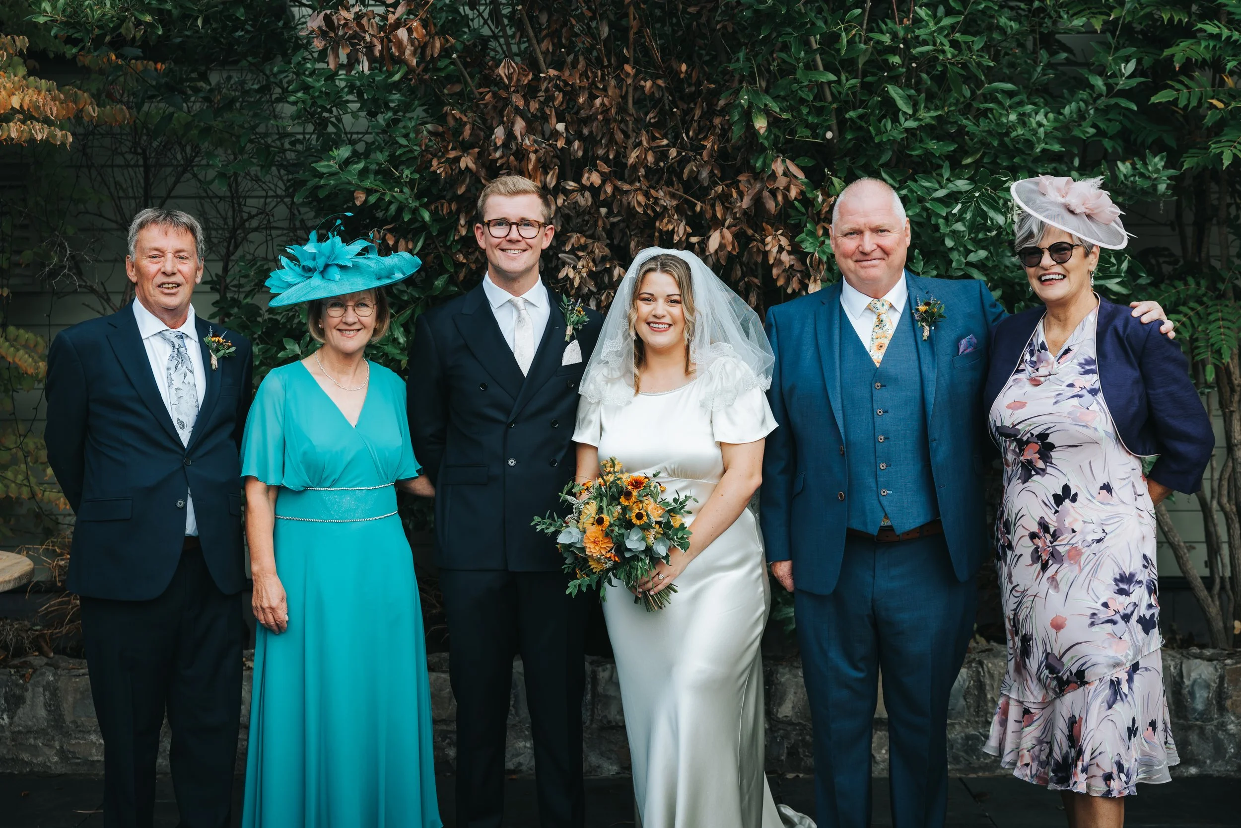 Group of wedding guests and a bride and groom standing outdoors in front of a leafy background, dressed in formal attire.