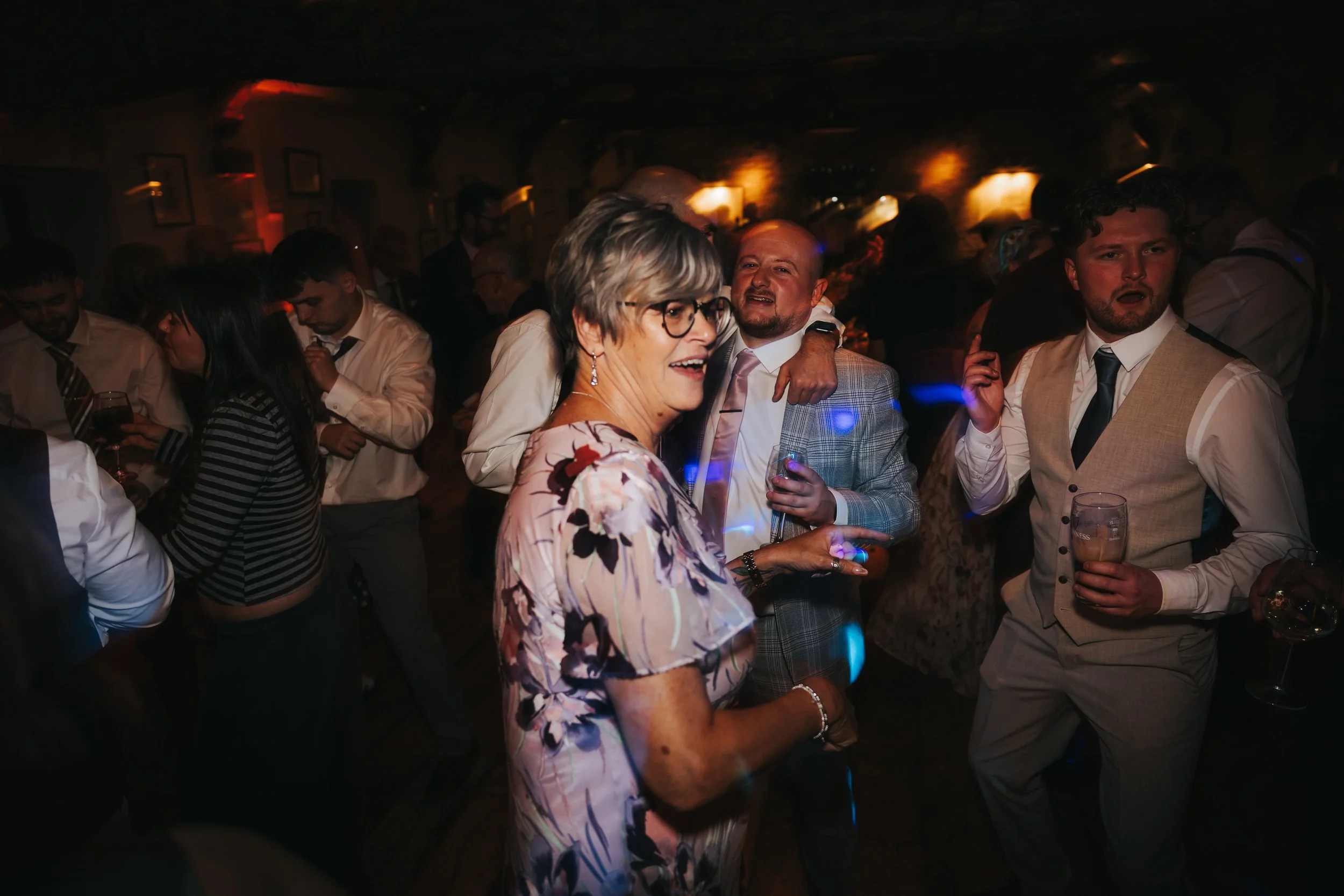 People dancing and socializing at a party or celebration in a dimly lit venue, with colorful lights.