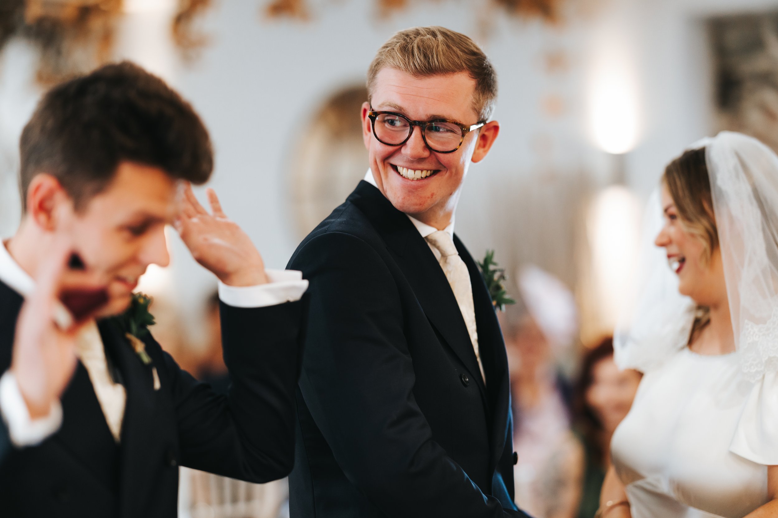 Groom smiling and looking at the bride during a wedding ceremony, with the officiant and best man nearby.
