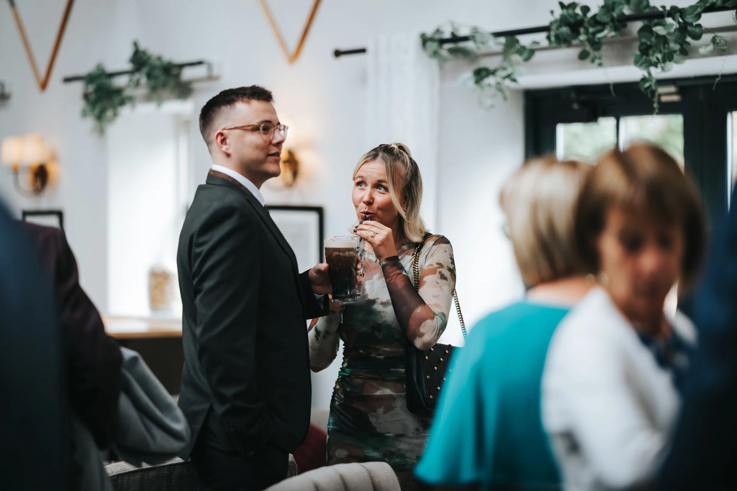 A man in a black suit holding a drink and a woman in a floral dress sipping a beverage with a straw at a social gathering in a decorated indoor venue.
