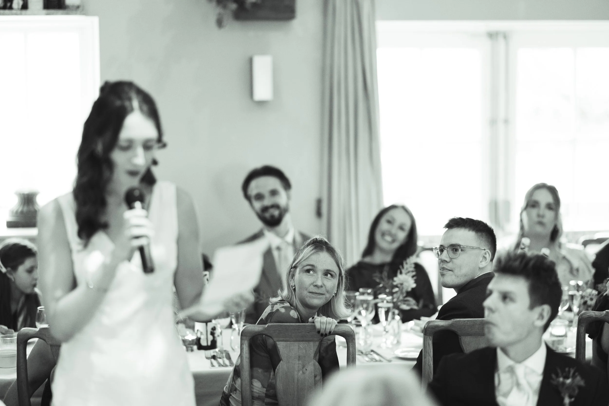 A woman giving a toast or speech with a microphone at a dining event, with guests seated at a table, some smiling and looking on, in a bright room with large windows.