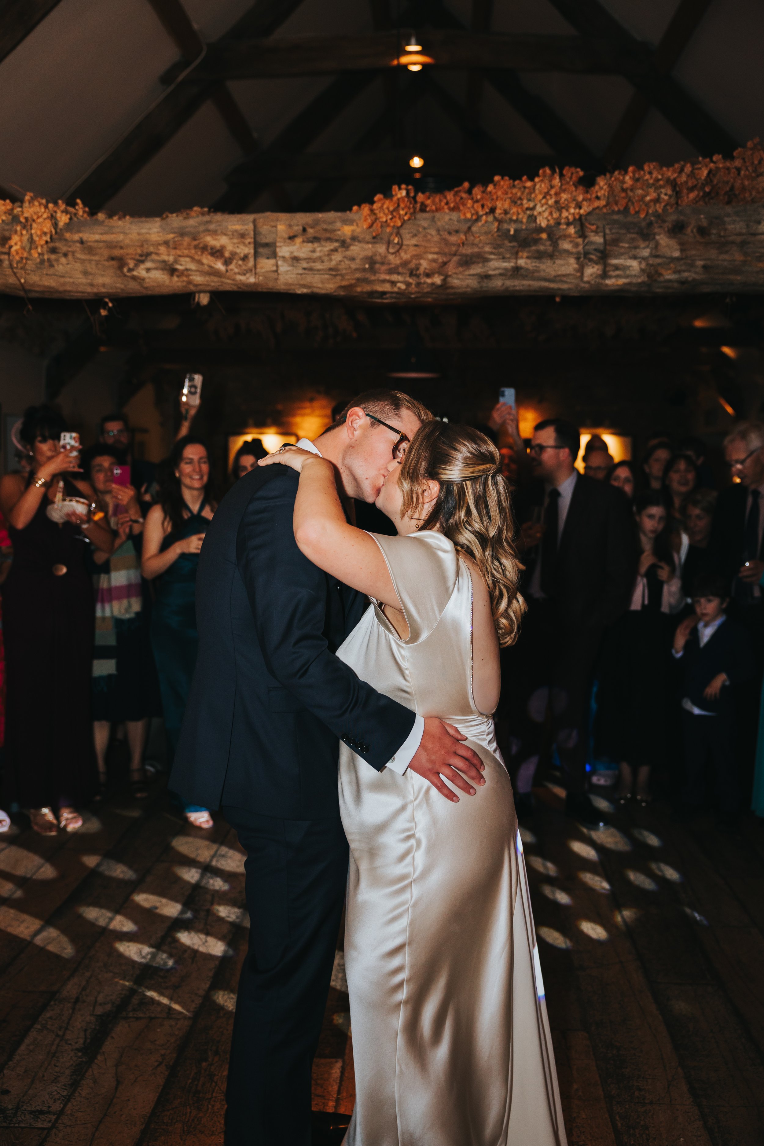 A bride and groom kissing during their wedding dance in a rustic barn setting with guests watching and taking photos.