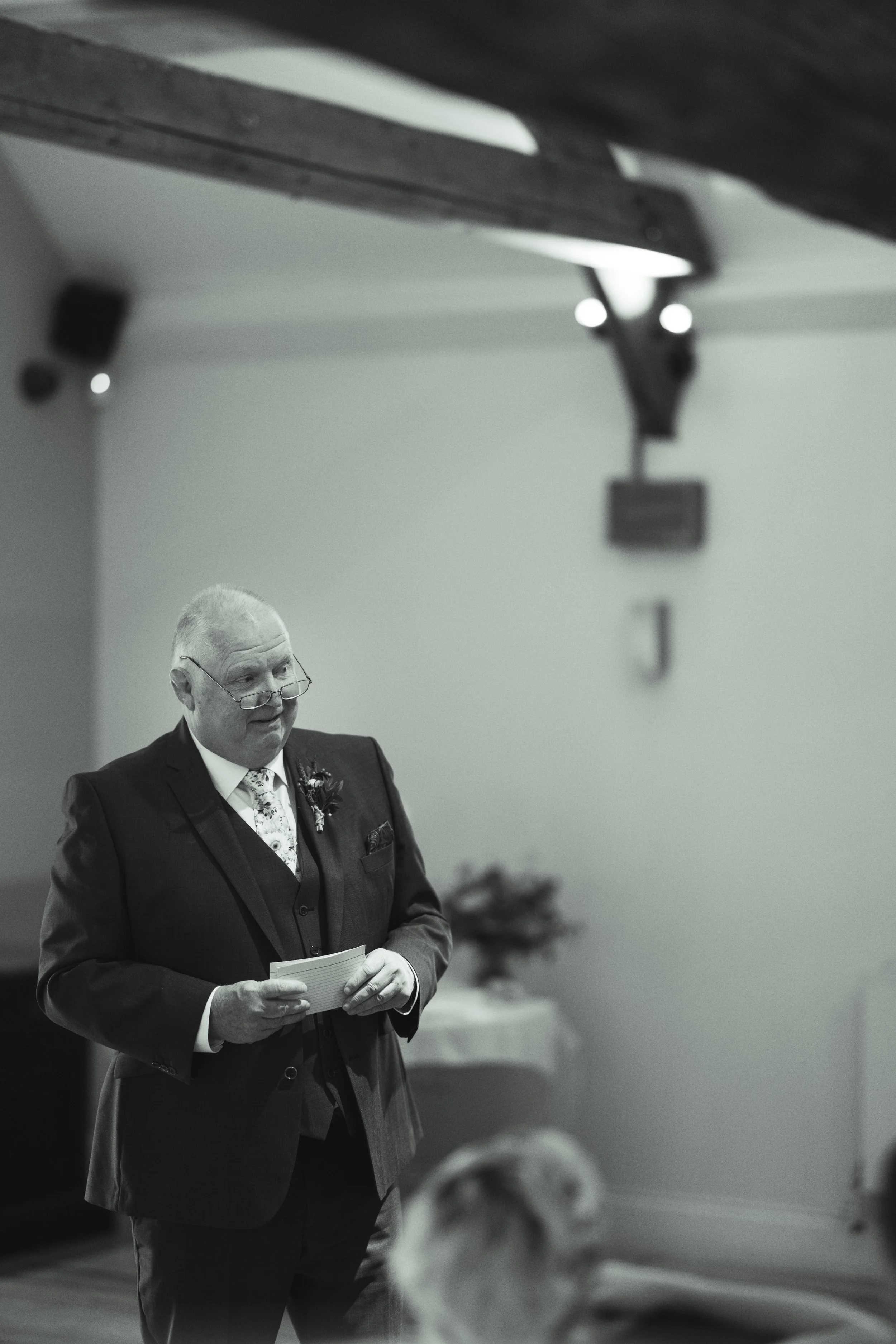 An elderly man dressed in a suit and tie, holding a piece of paper, standing indoors at a formal event or ceremony.