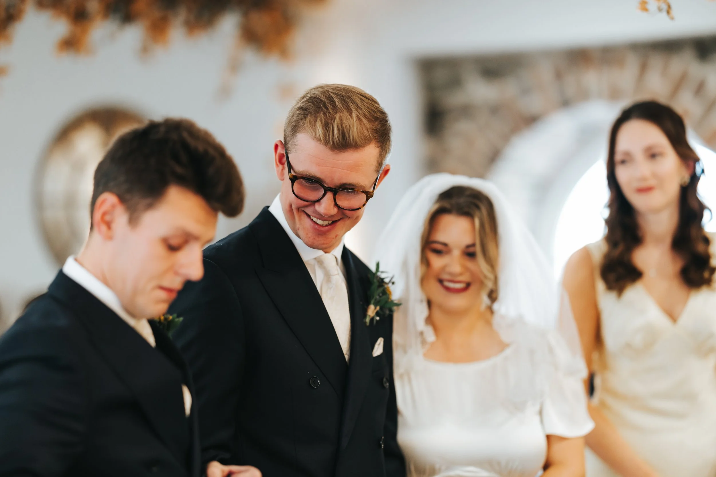 A wedding ceremony with four people, including a groom with glasses, a bride, and two others, all smiling and dressed in formal attire, standing inside a room with stone walls and large windows.