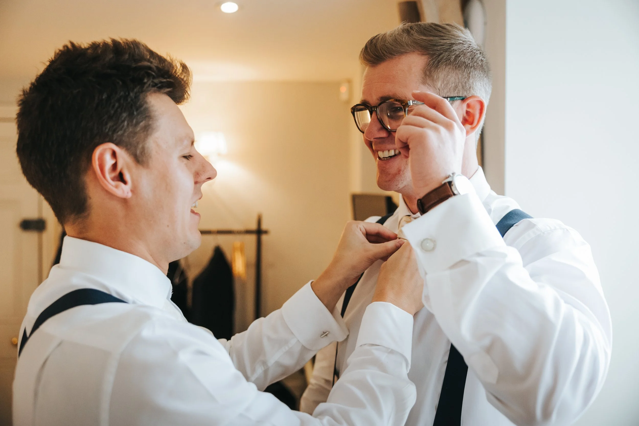 Two men in white shirts and black suspenders smiling as one helps the other adjust his bow tie in a warmly lit room.