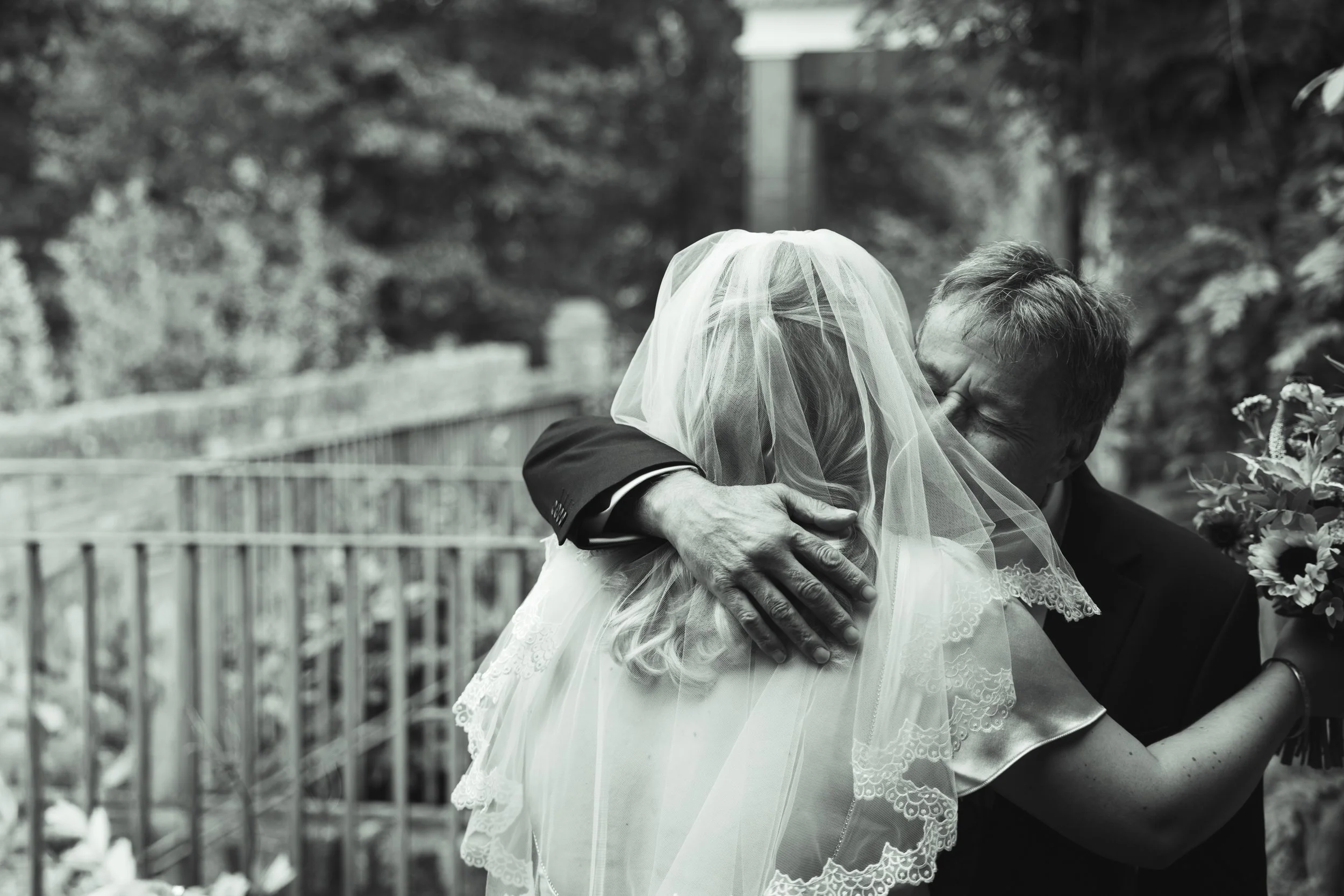 A joyful woman wearing a wedding veil and dress hugging an older man in a tuxedo, both with their eyes closed, outdoors with trees and fencing in the background.