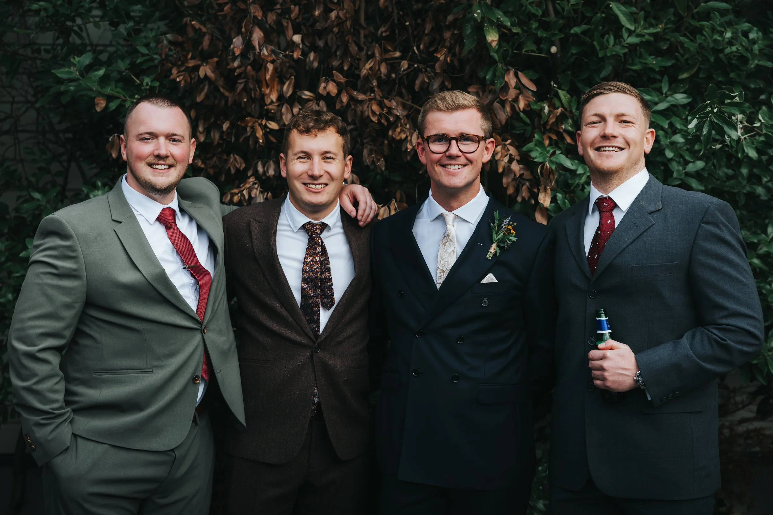 Five men in suits smiling and posing outdoors in front of a leafy background, with one man in the middle wearing a boutonniere.