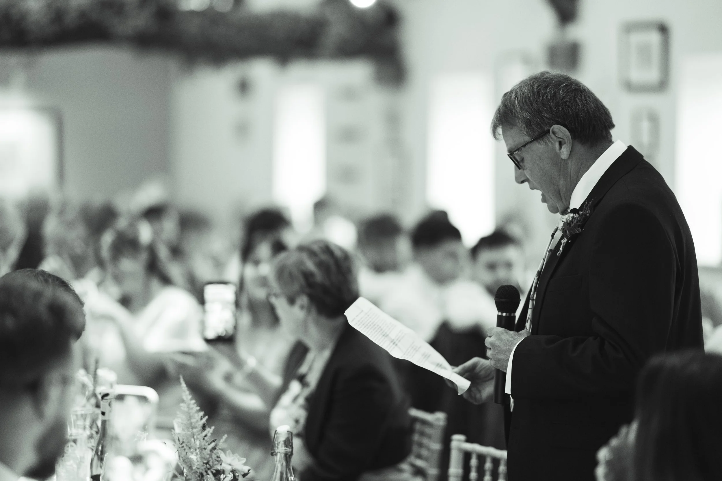 A man in a suit holding a microphone and a piece of paper, speaking at a formal event with seated guests listening.