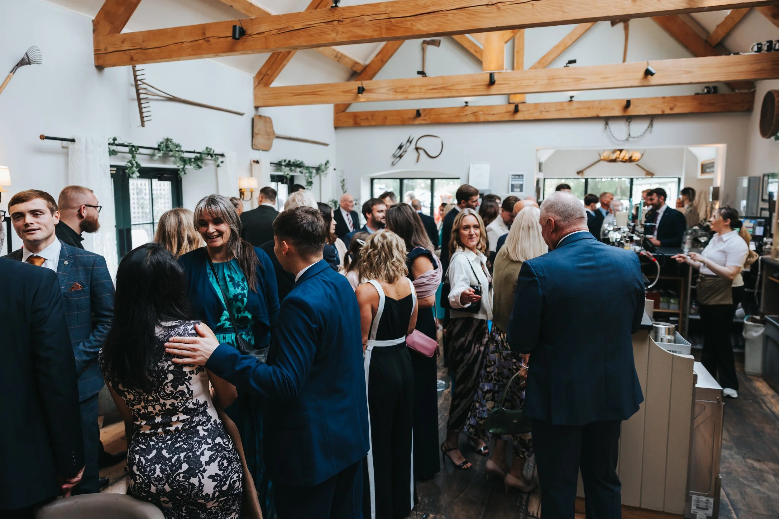 People socializing at a crowded indoor event with a bar counter on the right, wooden ceiling beams, and rustic decor.