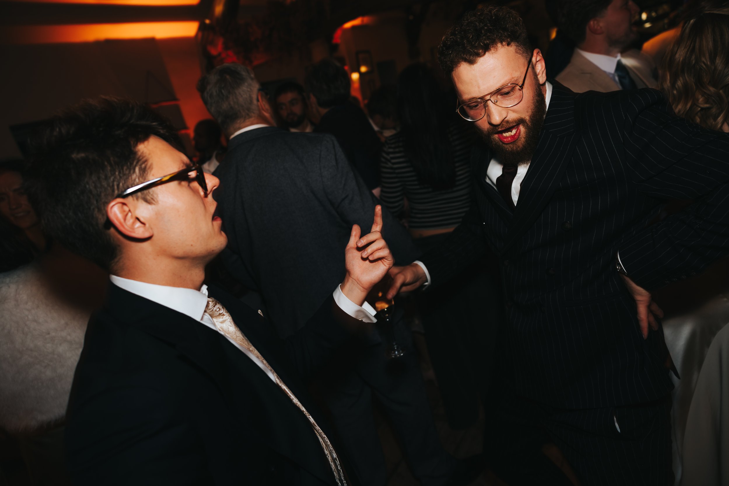 Two men in suits and glasses dancing and talking at a lively indoor social event with warm lighting and a crowd in the background.
