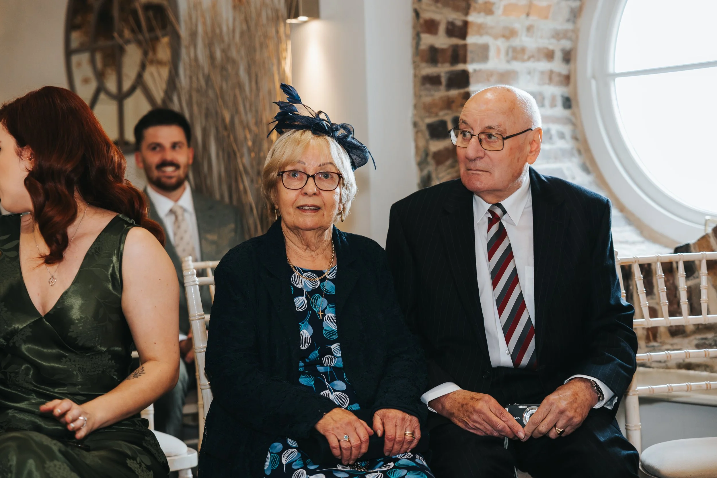 An elderly woman with glasses and a blue hat sitting next to an elderly man with glasses, both dressed formally, at a social event in a room with exposed brick walls and a large round window.
