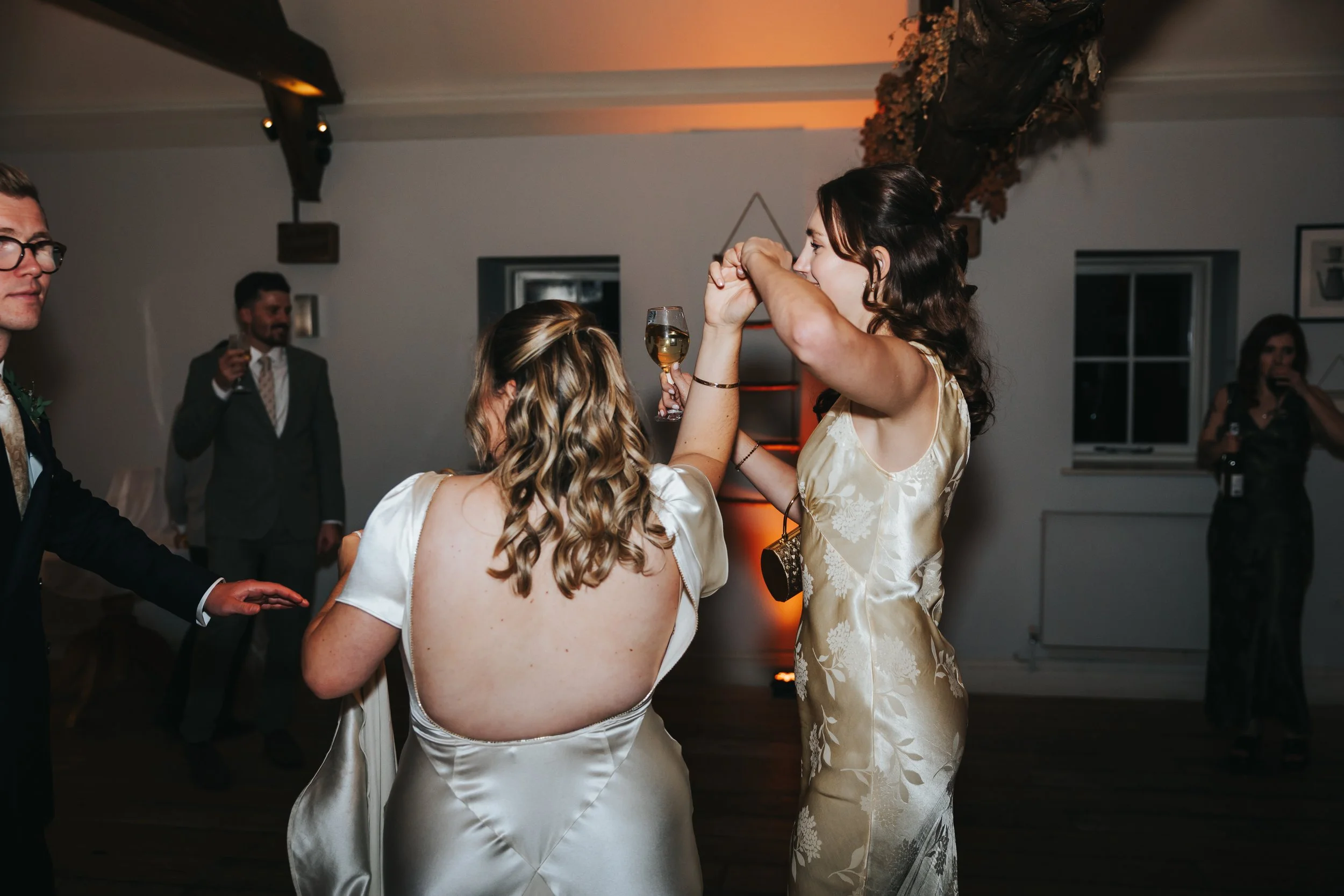 Two women dancing and clinking glasses of white wine at a wedding reception, with three men in suits in the background and a woman taking photos.