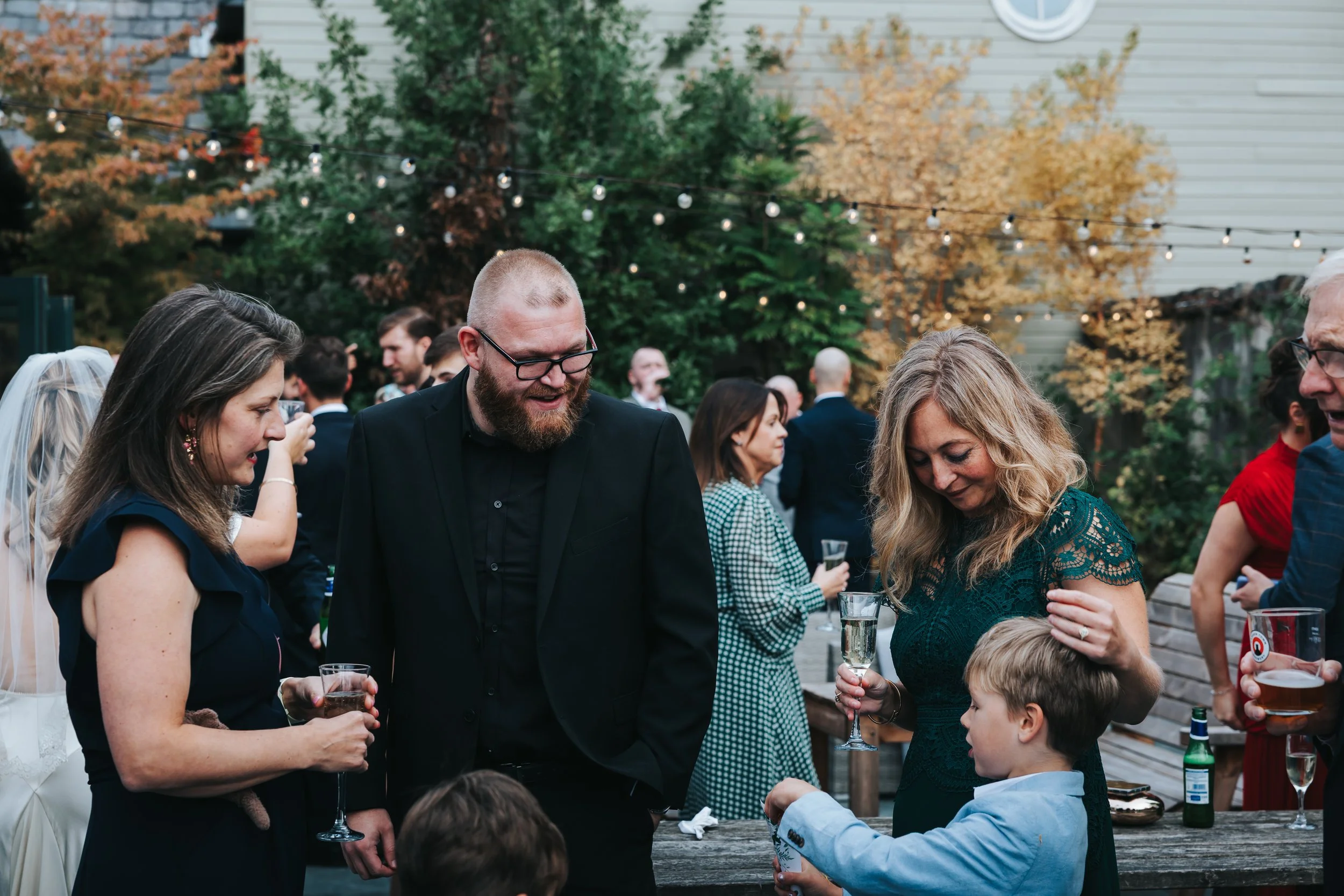 People socializing at an outdoor gathering during the evening, with string lights and autumn trees in the background. A woman in a green lace dress and a woman in a dark dress are talking to children, with others holding drinks nearby.