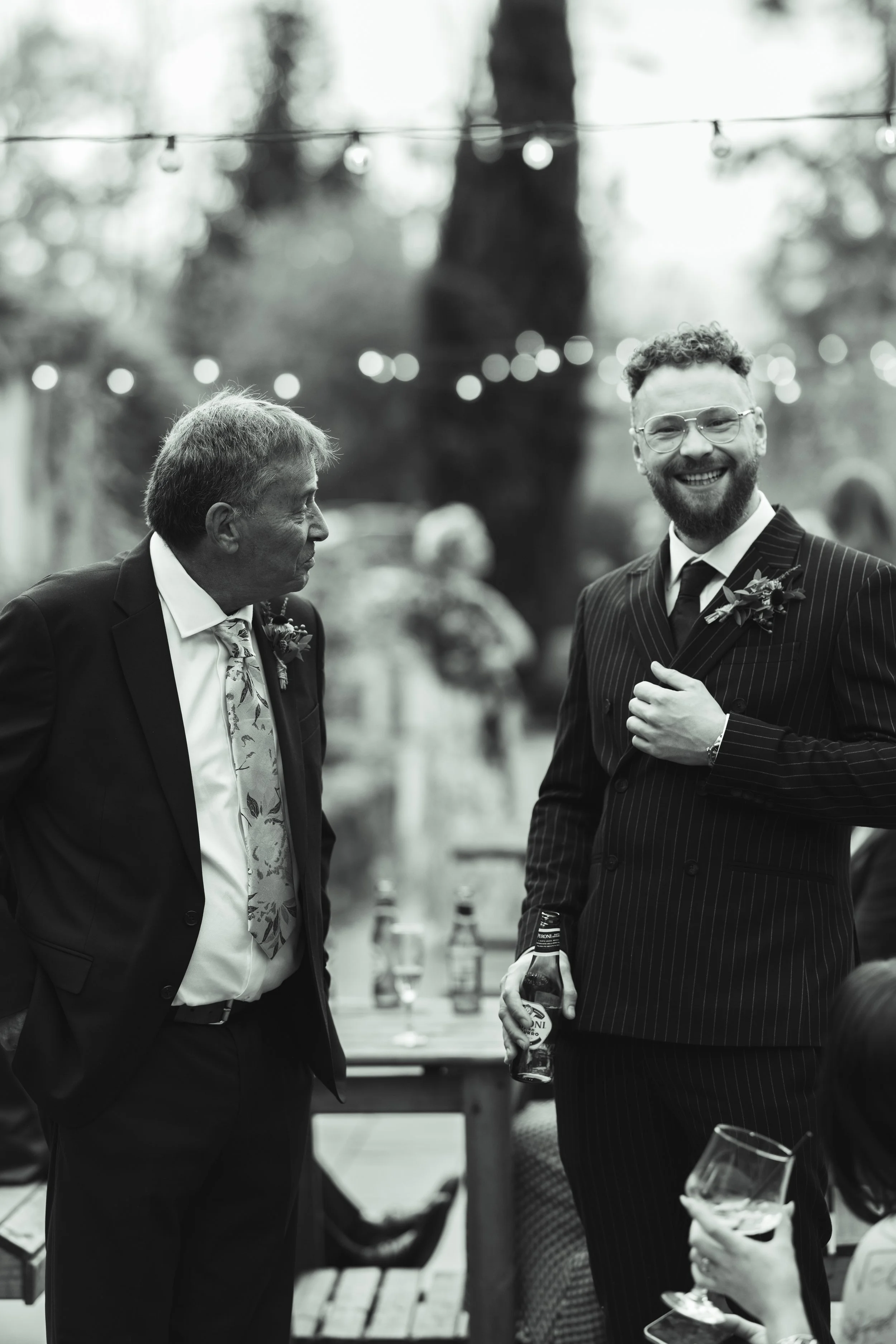 Two men dressed in suits at an outdoor gathering, engaging in conversation, with one holding a beer bottle and the other a glass of wine, with string lights and other guests in the background.