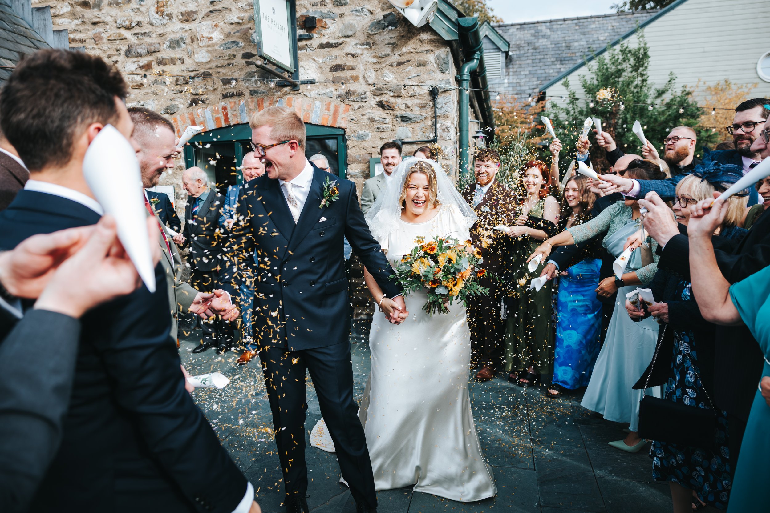 A newlywed couple holding hands and smiling, surrounded by a crowd of guests cheering and throwing confetti during an outdoor wedding celebration.