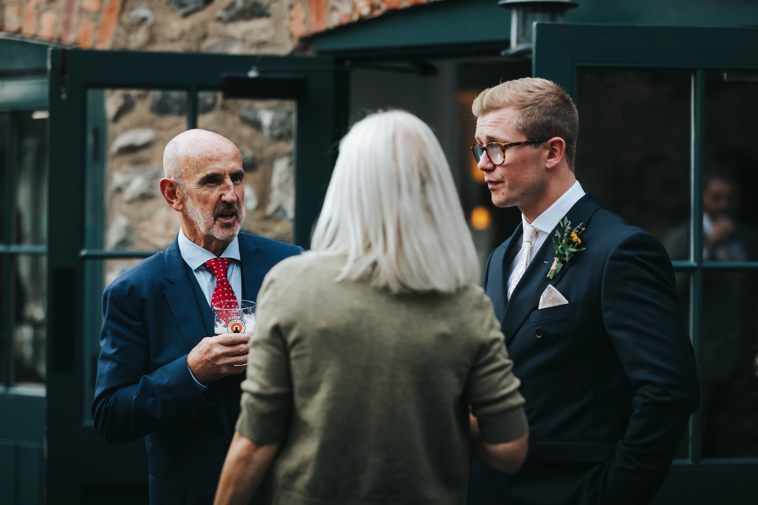 Three people in formal attire engaged in conversation at an outdoor event, with a stone wall and window behind them.