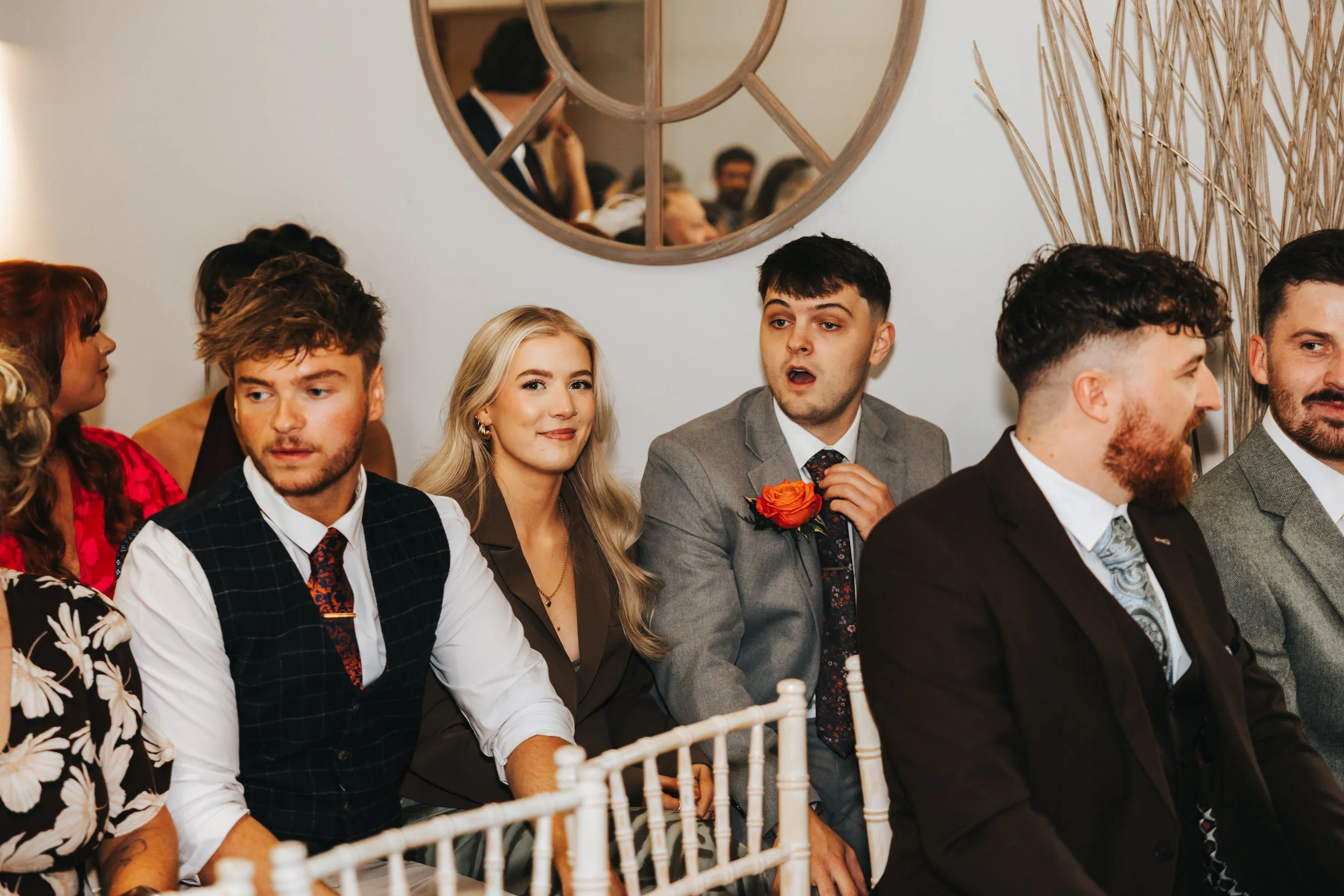 Group of people sitting and listening at what appears to be a formal event or wedding, with a mirror reflecting others in the background.
