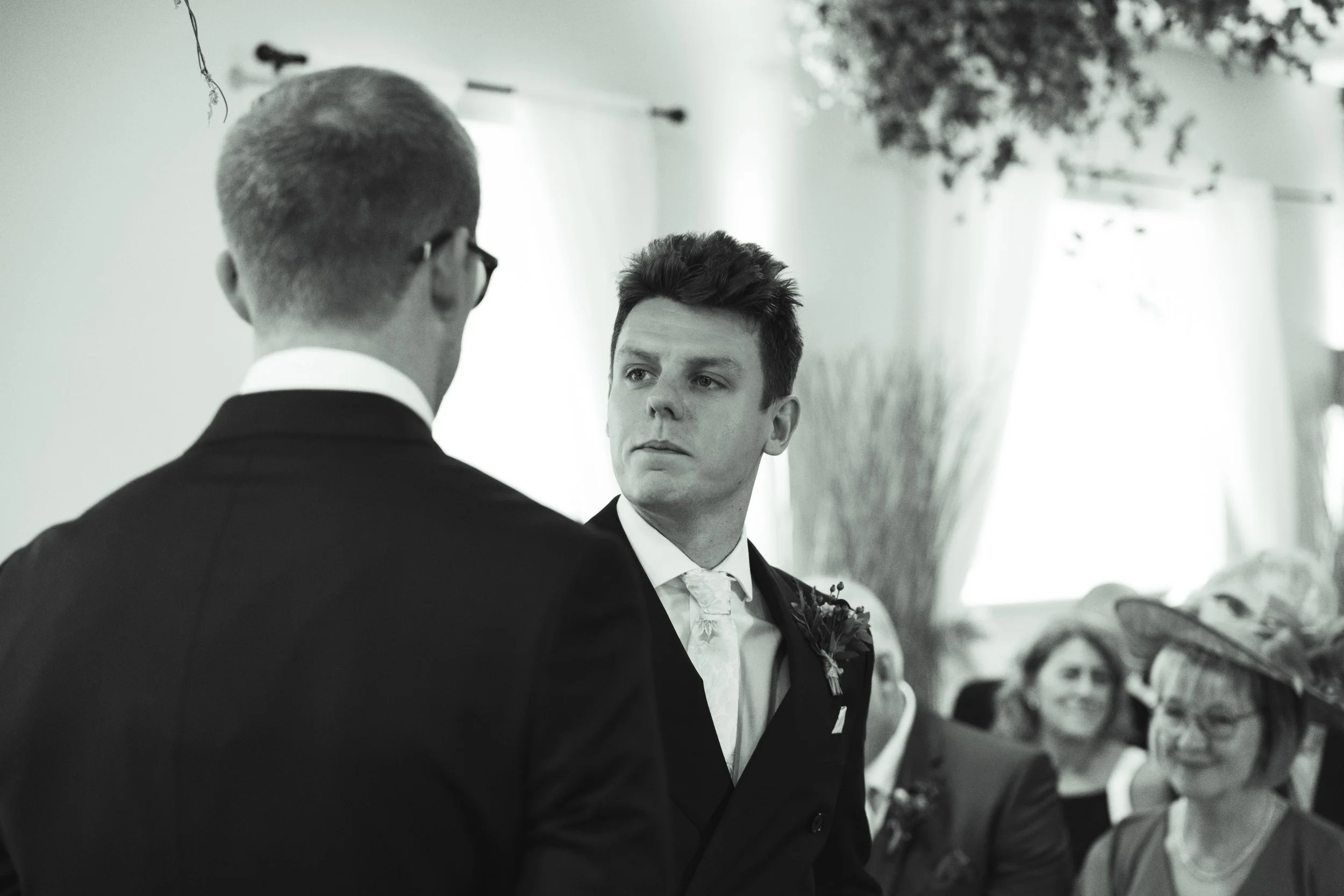 Black and white photo of two men facing each other during a formal event, possibly a wedding, with women in the background. One man is wearing glasses and a dark suit, and the other man is also in a suit, with a serious expression.