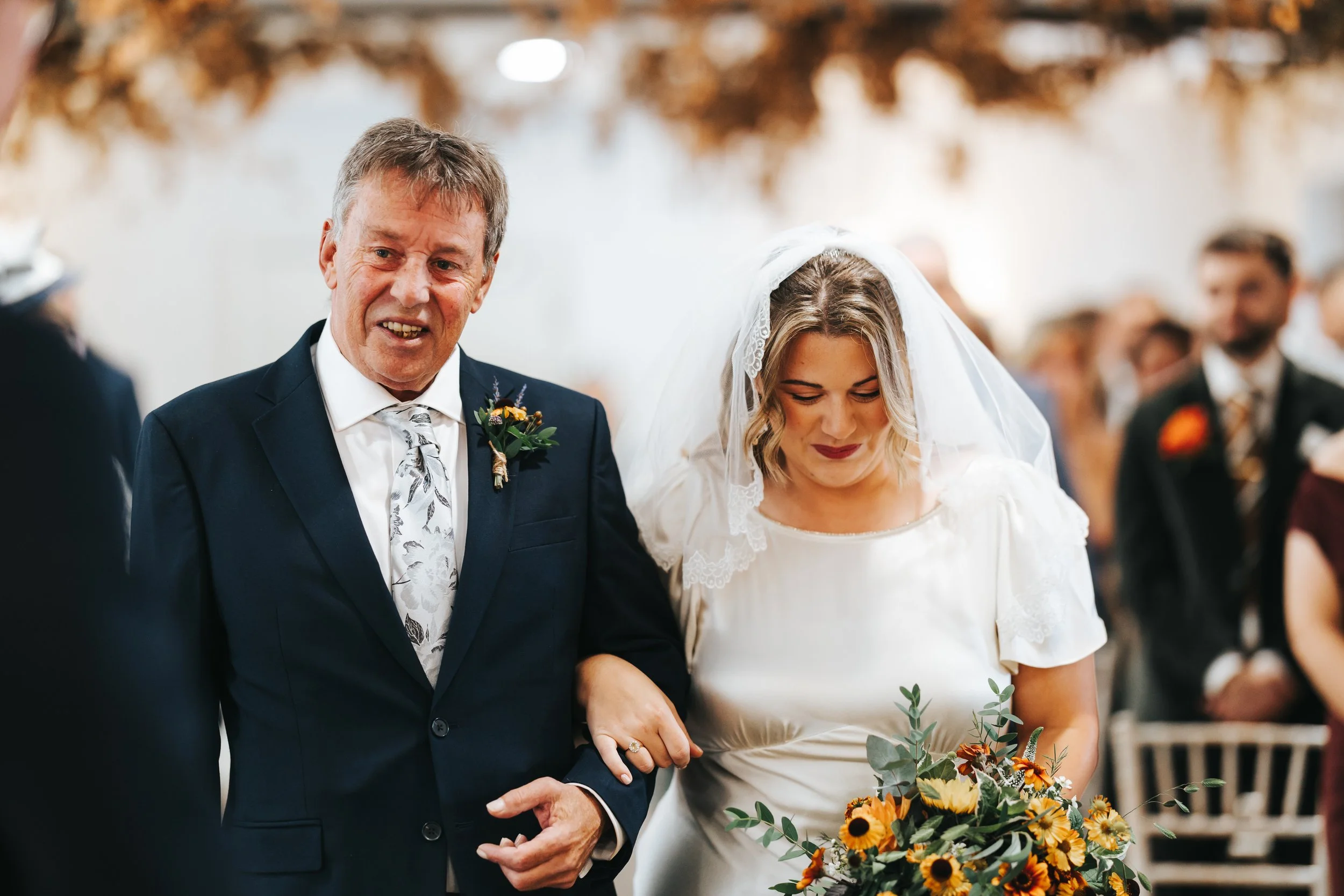 A bride and an older man walking down the aisle at a wedding ceremony. The bride is holding a bouquet of sunflowers and greenery, and the man is dressed in a dark suit with a boutonniere.