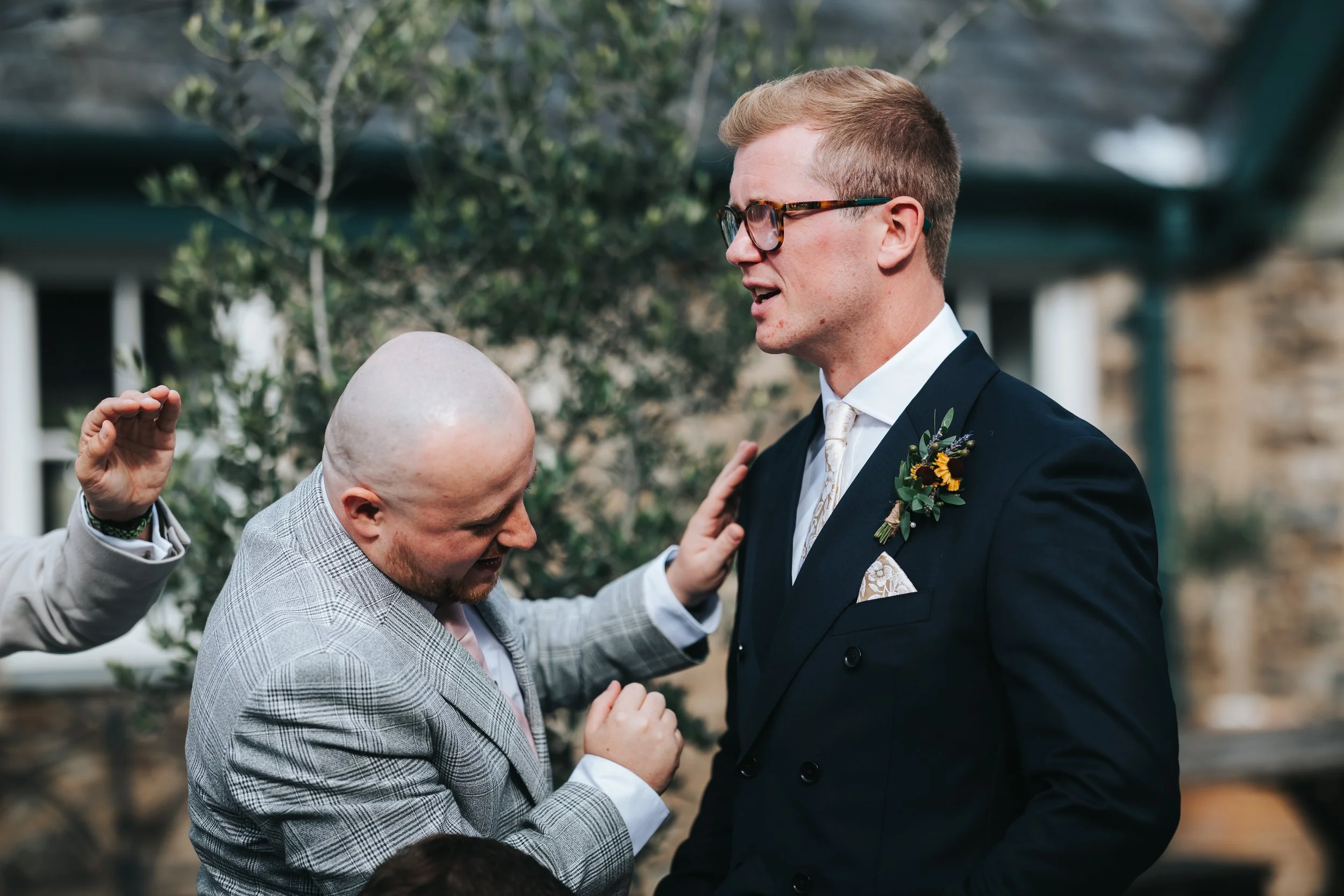 A man with glasses, wearing a dark suit and a boutonniere, is outdoors during a wedding ceremony. Another man with a bald head and checked suit appears to be emotional, with someone else raising a hand nearby.