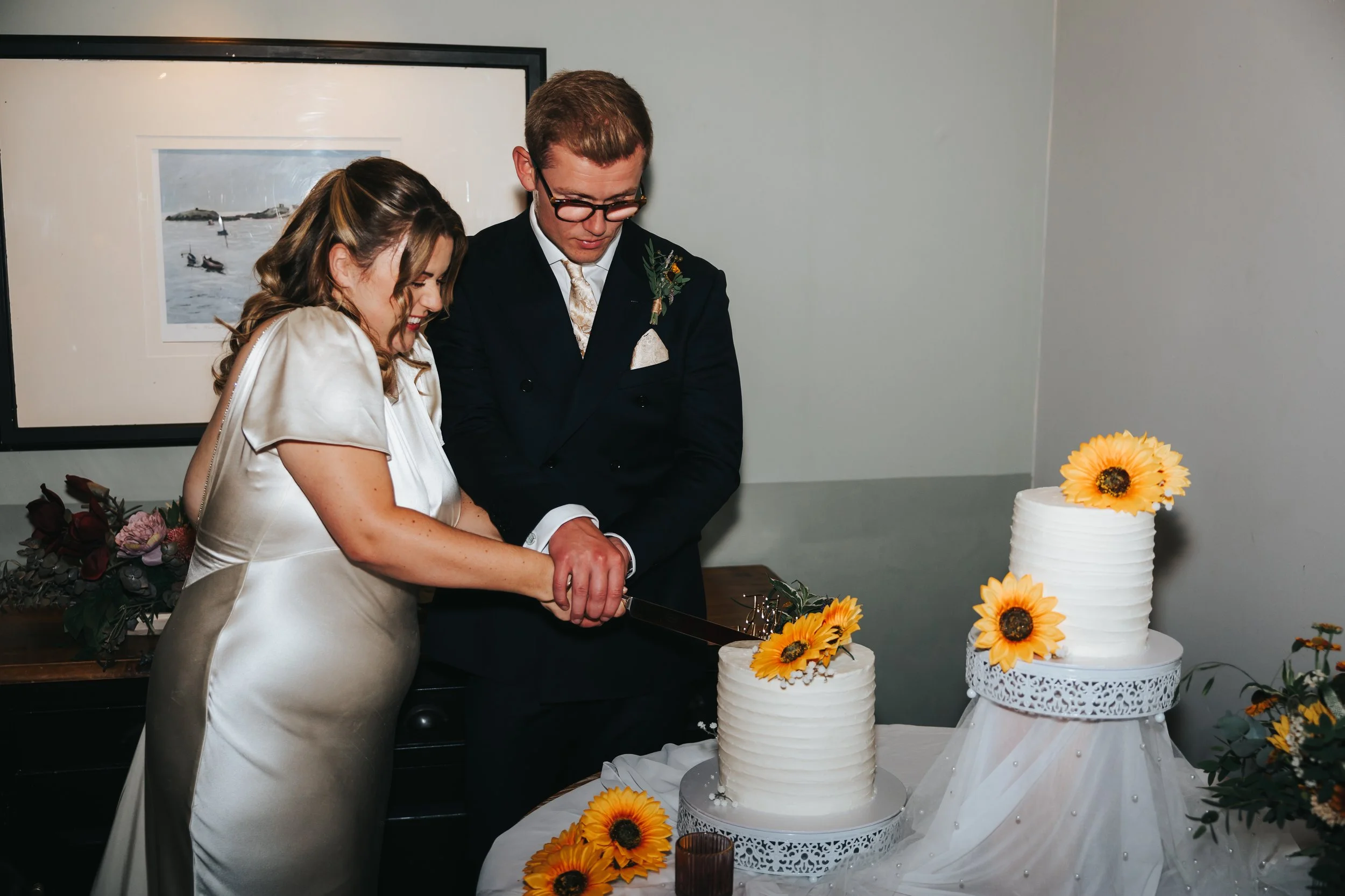 A couple in wedding attire cut a wedding cake decorated with sunflower flowers. The bride has light brown hair styled in loose curls and is wearing a satin wedding dress. The groom has short hair, glasses, and is dressed in a black suit with a bouton