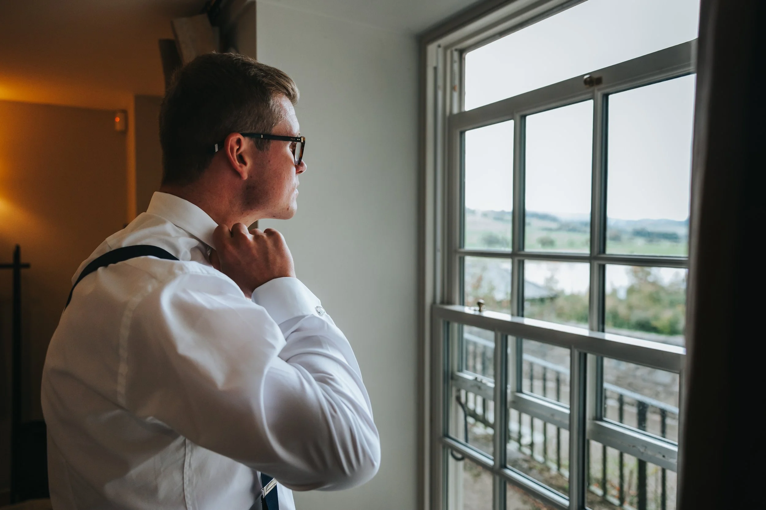 A man in a white shirt and tie, wearing glasses, looking out a window and adjusting his collar.