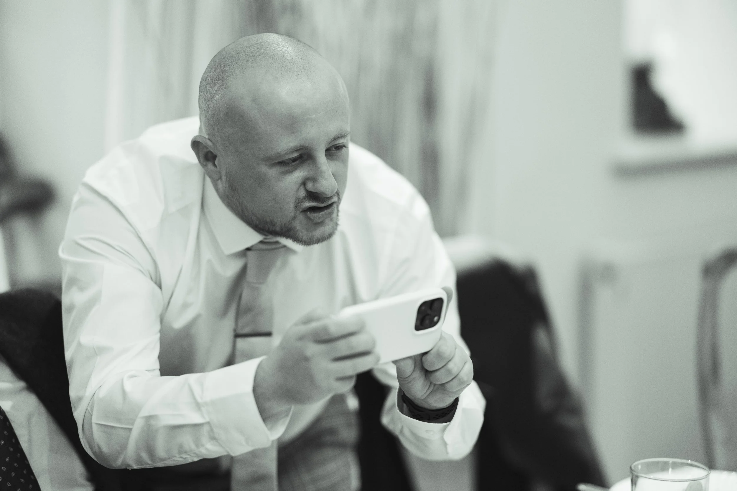A man with a shaved head and white shirt holding a smartphone in a room, possibly taking a photo or video.