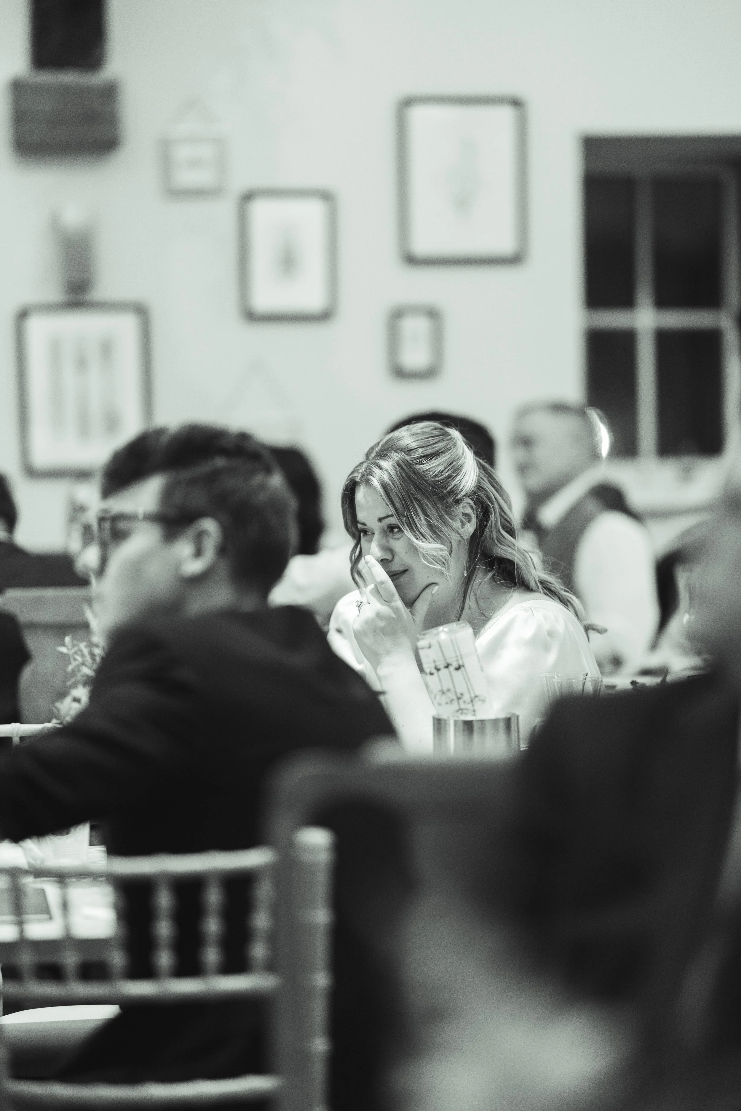 A woman with wavy hair and a white shirt sitting at a table, resting her chin on her hand, in a dimly lit room with framed pictures on the wall, during a social gathering.